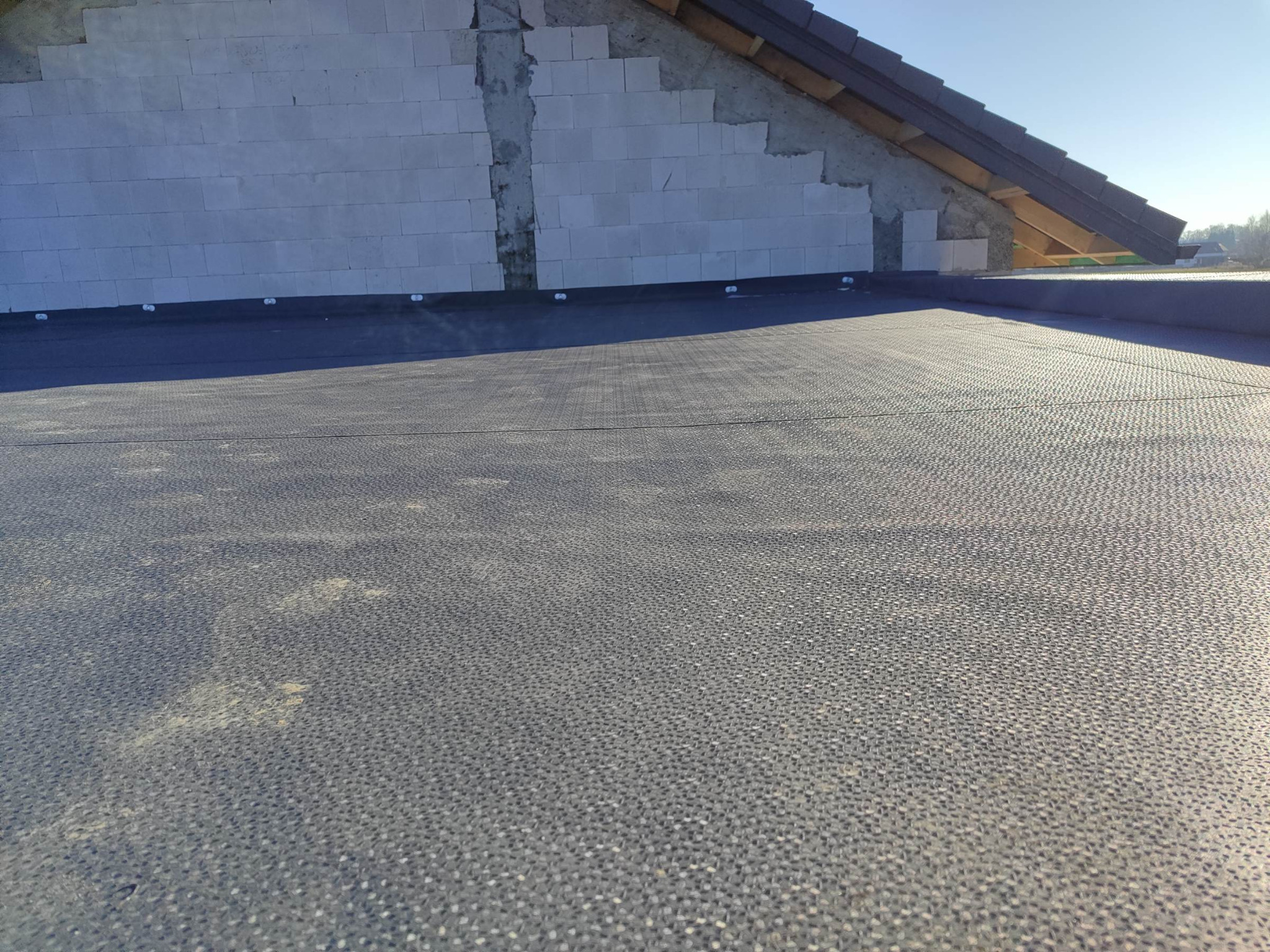 Czarna membrana dachowa na płaskim dachu z widoczną ścianą budynku w trakcie budowy. Widok z góry na pokrycie dachowe.