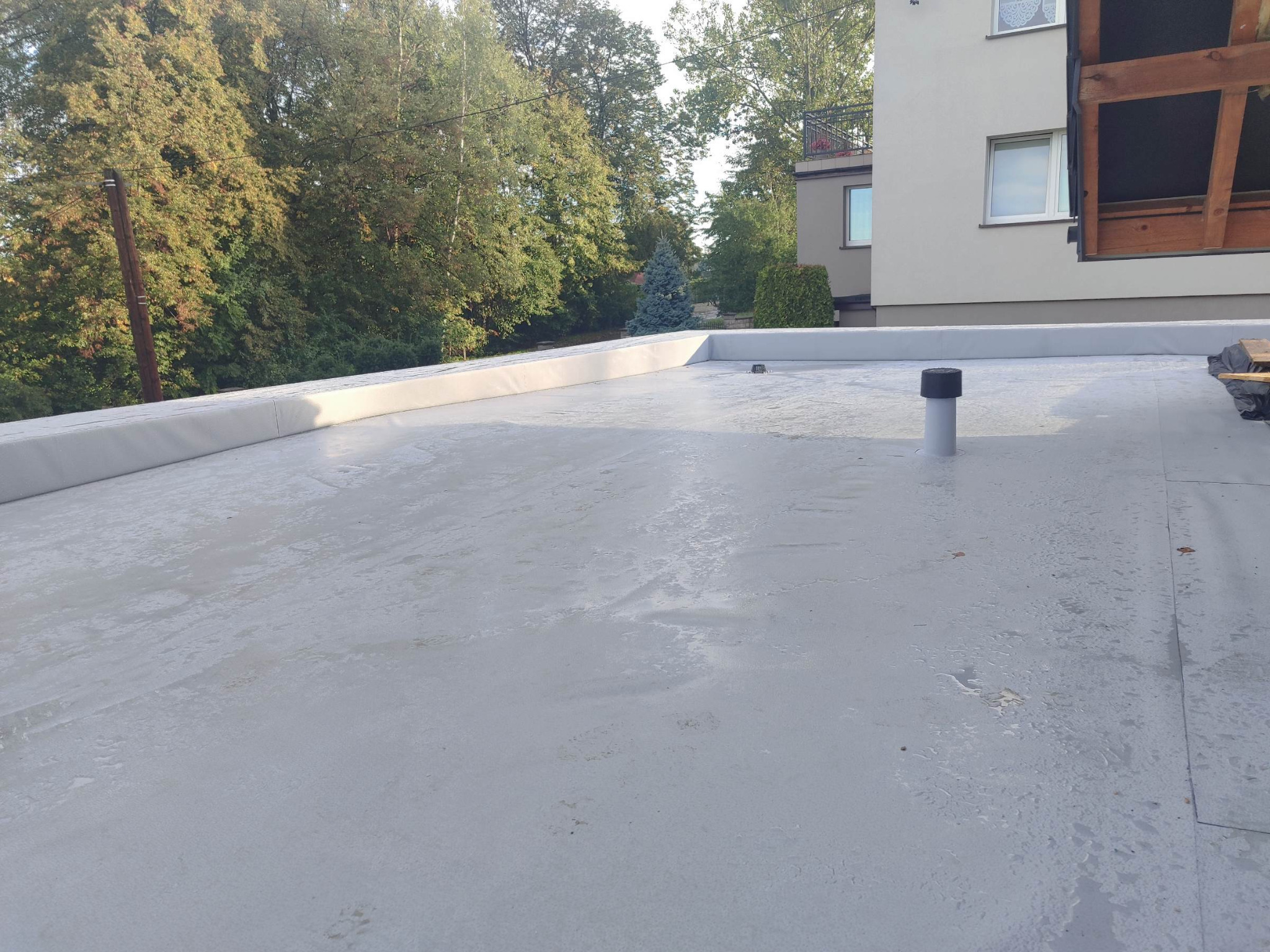 Jasnoszary, płaski dach po renowacji z kominkiem wentylacyjnym na tle zieleni i budynku. Widoczne detale wykończenia krawędzi dachu.