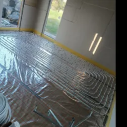 Rozłożone rury ogrzewania podłogowego na folii z czerwoną siatką w surowym wnętrzu z oknami i częściowo wykończonymi ścianami z płyt gipsowo-kartonowych. Widoczne narzędzia montażowe i zwój rur.