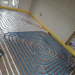 Grzegorz Kaczmarek - Wnętrze w trakcie montażu ogrzewania podłogowego: białe i niebieskie rury ułożone spiralnie na folii izolacyjnej z czerwoną siatką, przytwierdzone plastikowymi uchwytami, widoczne surowe ściany...