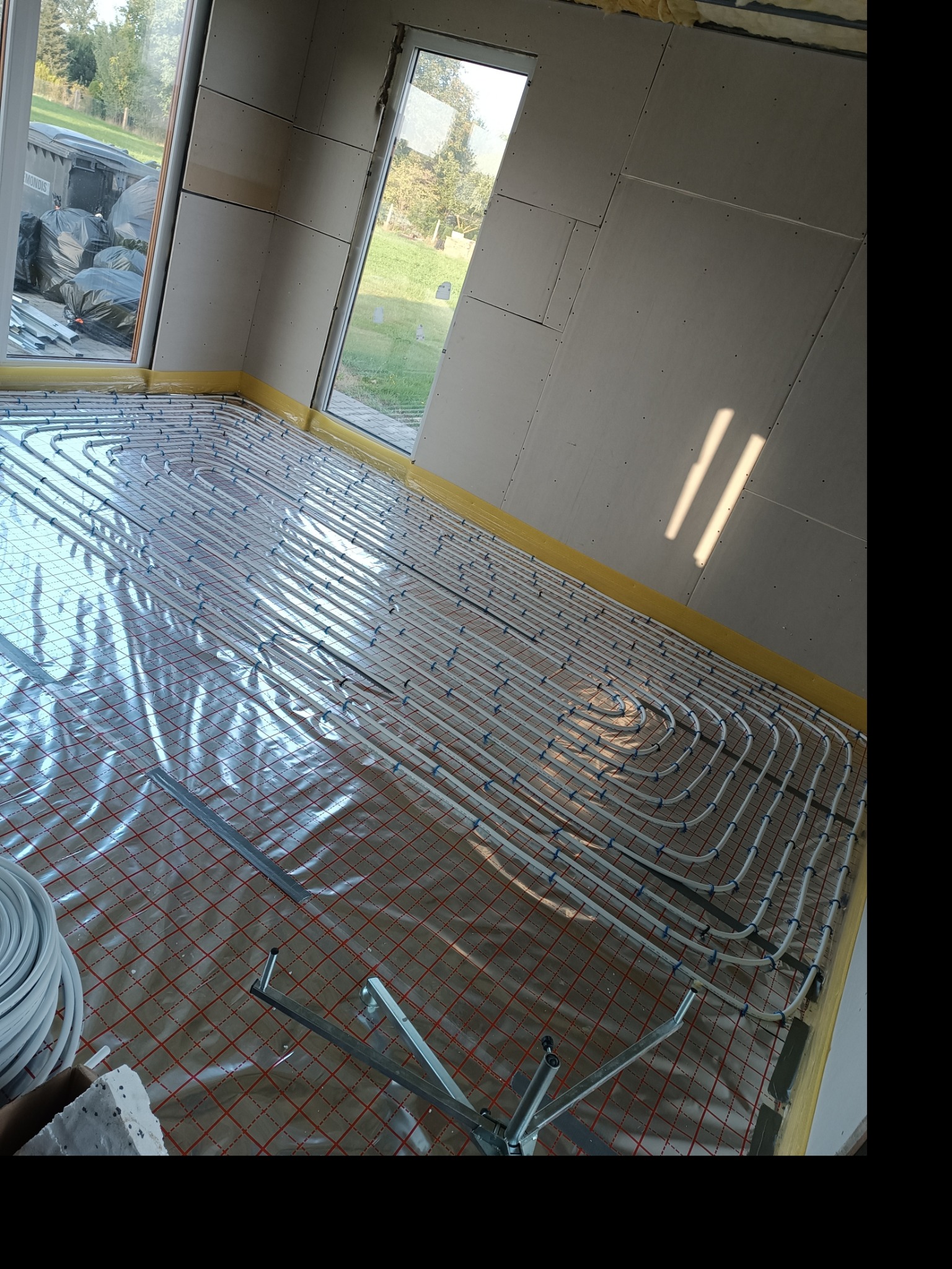 Rozłożone rury ogrzewania podłogowego na folii z czerwoną siatką w surowym wnętrzu z oknami i częściowo wykończonymi ścianami z płyt gipsowo-kartonowych. Widoczne narzędzia montażowe i zwój rur.