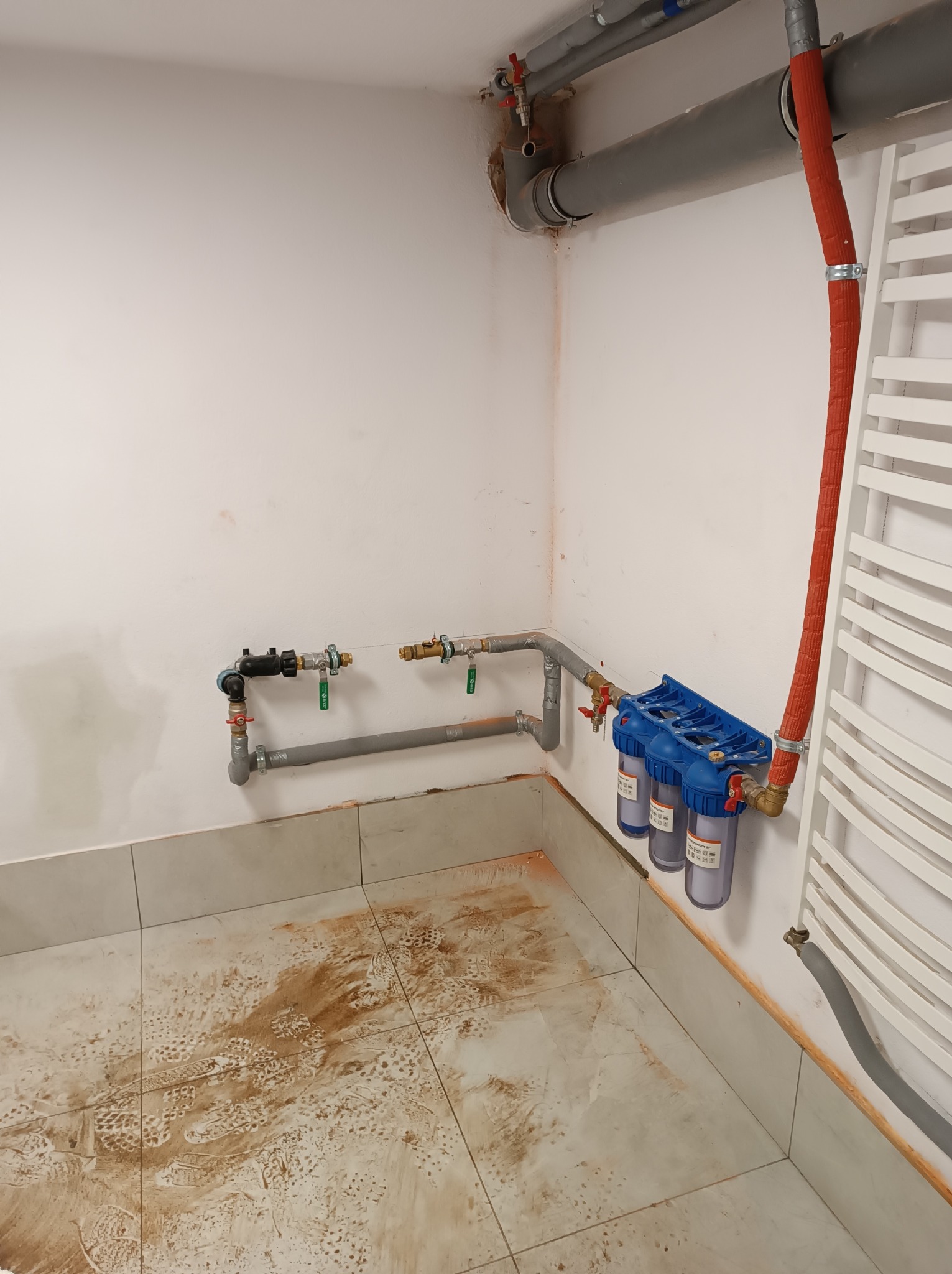 Fragment łazienki w trakcie prac hydraulicznych: widoczne rury, filtry wody z niebieską obudową, grzejnik drabinkowy oraz zabrudzona podłoga z widocznymi śladami butów.