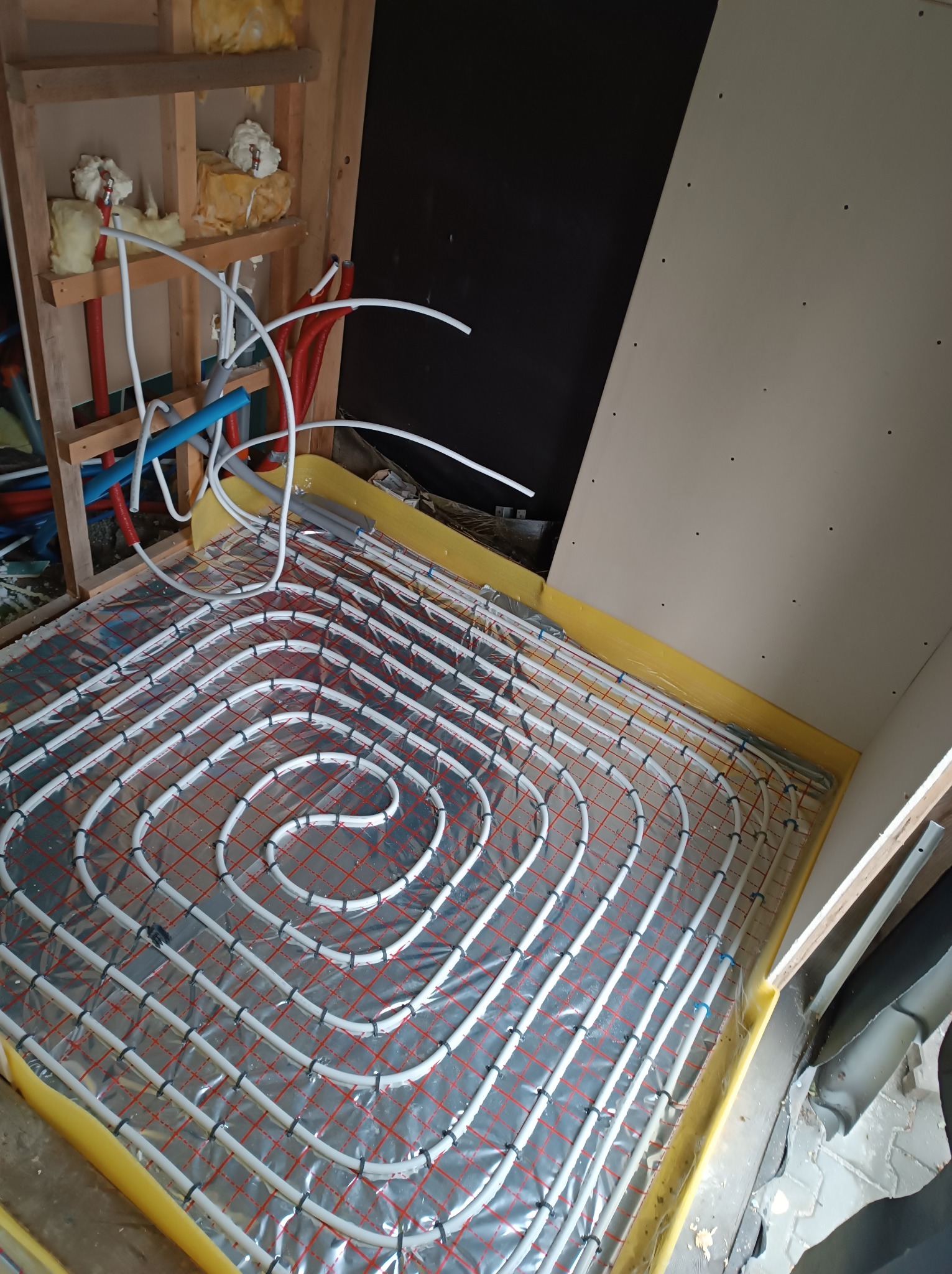 Układanie rur ogrzewania podłogowego na folii z siatką, widoczne fragmenty ścian w surowym stanie i wystające rury instalacji.