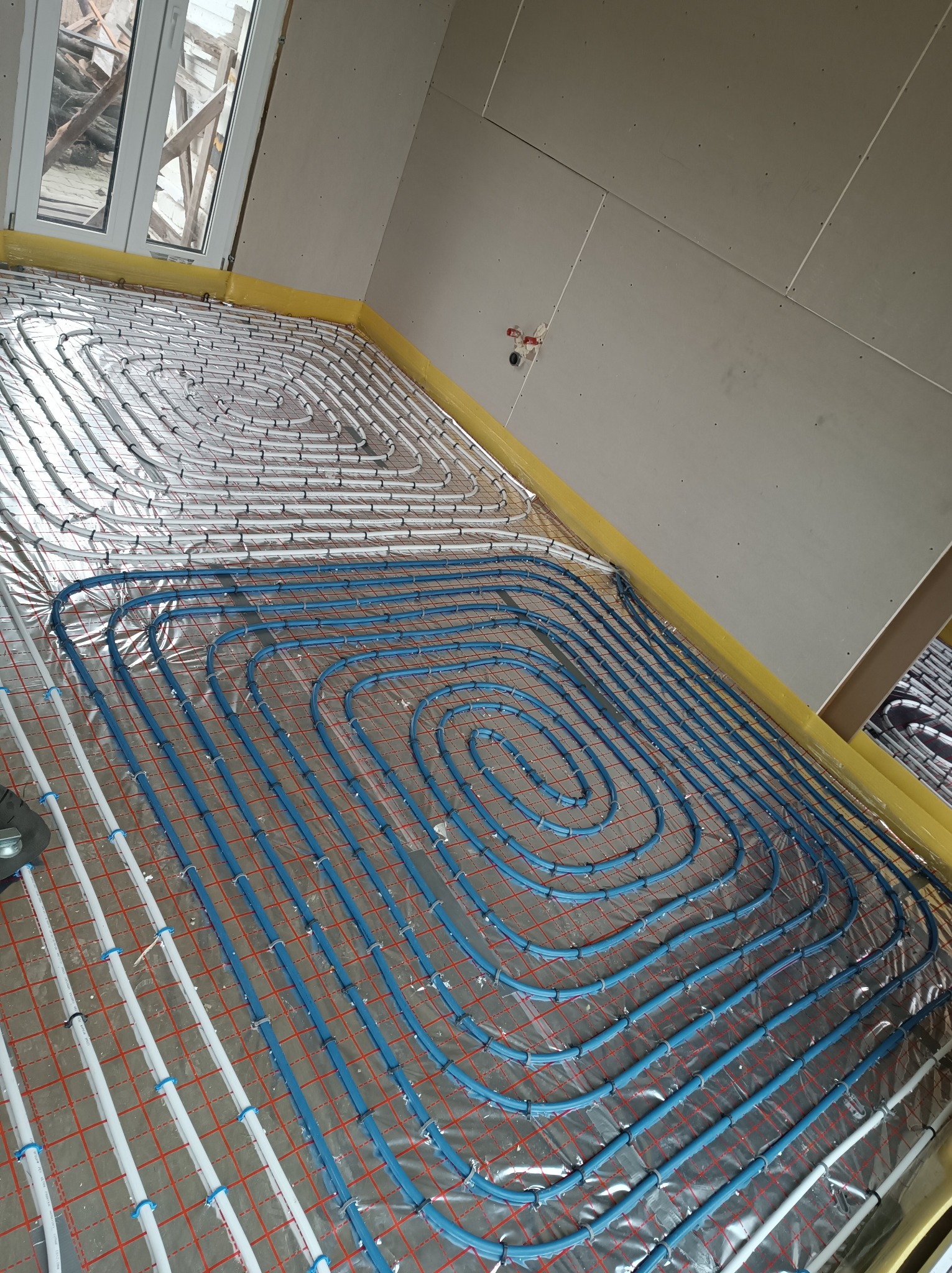 Wnętrze w trakcie montażu ogrzewania podłogowego: białe i niebieskie rury ułożone spiralnie na folii izolacyjnej z czerwoną siatką, przytwierdzone plastikowymi uchwytami, widoczne surowe ściany...