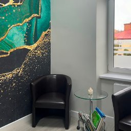 Studio Stomatologii Estetycznej DENTIME Jarosław - Poczekalnia z dwoma skórzanymi fotelami, stolikiem z czasopismami i dekoracyjną tapetą w odcieniach zieleni i złota. Okno z widokiem na budynek z czerwonym dachem.