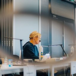 Studio Stomatologii Estetycznej DENTIME Jarosław - Protetyczka w okularach powiększających, pracująca przy biurku z lampą i narzędziami, w tle widoczne okna z żaluzjami.