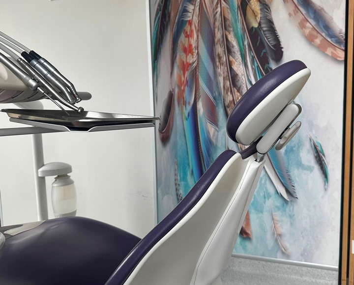 Fotel dentystyczny z narzędziami na tacy, na tle ściany z dekoracyjnym obrazem piór.