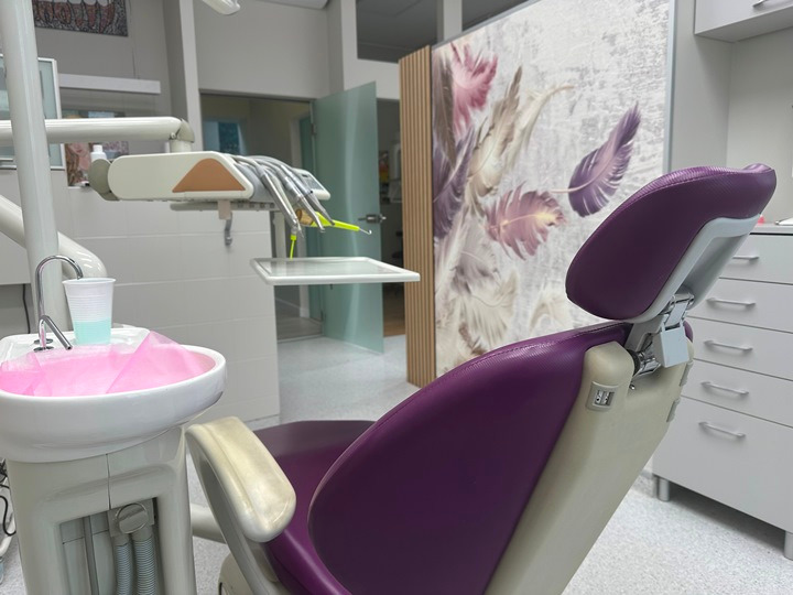 Fioletowy fotel dentystyczny w gabinecie z umywalką, narzędziami i dekoracyjnym parawanem w tle.