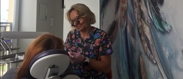 Lekarz w okularach lupowych i rękawiczkach, w gabinecie, podczas badania pacjenta przy fotelu dentystycznym; w tle ściana z motywem piór.