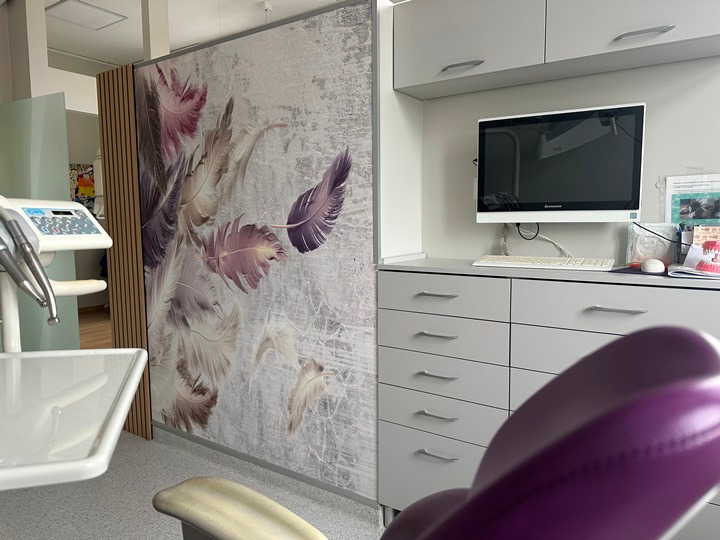 Wnętrze gabinetu stomatologicznego z nowoczesnym wyposażeniem, urządzeniem do zabiegów, biurkiem z komputerem i ścianą ozdobioną motywem piór