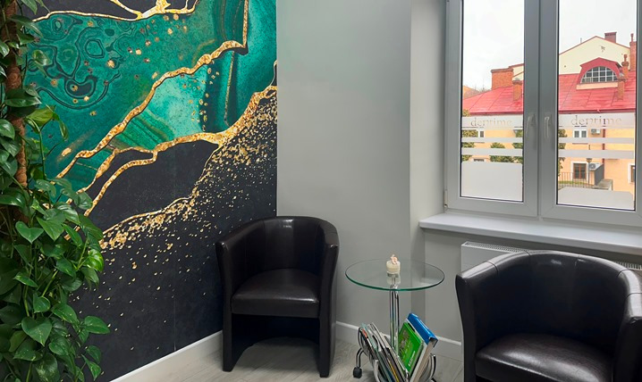 Poczekalnia z dwoma skórzanymi fotelami, stolikiem z czasopismami i dekoracyjną tapetą w odcieniach zieleni i złota. Okno z widokiem na budynek z czerwonym dachem.