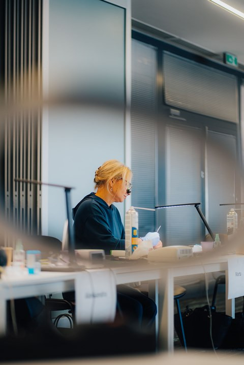 Protetyczka w okularach powiększających, pracująca przy biurku z lampą i narzędziami, w tle widoczne okna z żaluzjami.