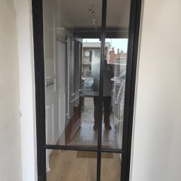 Metalwork - Czarna, przeszklona konstrukcja drzwiowa z podziałami, widoczna z perspektywy wnętrza, z odbiciem osoby robiącej zdjęcie na szybie. W tle widoczne elementy wyposażenia wnętrza oraz ulica.