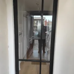 Czarna, przeszklona konstrukcja drzwiowa z podziałami, widoczna z perspektywy wnętrza, z odbiciem osoby robiącej zdjęcie na szybie. W tle widoczne elementy wyposażenia wnętrza oraz ulica.