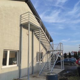 Metalwork - Zewnętrzne, metalowe schody ewakuacyjne przy jasnym budynku z dwoma oknami, widoczne z boku, z beczką w tle i fragmentem samochodu oraz niebem z chmurami.