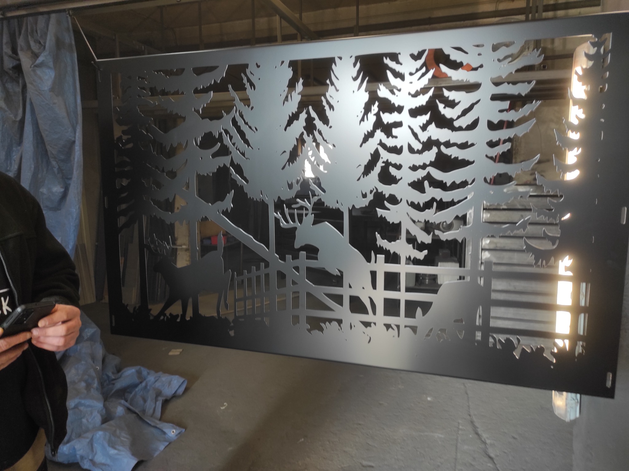 Panel metalowy z wycinanym laserowo motywem leśnym, przedstawiającym jelenie i drzewa, trzymany przez osobę z telefonem w dłoni, na tle warsztatu.