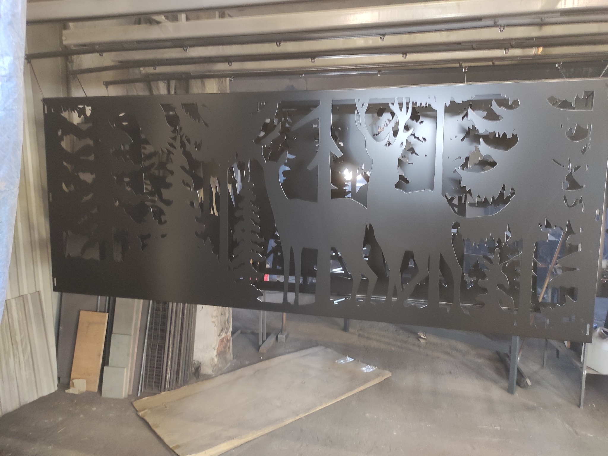 Czarny, metalowy panel dekoracyjny z wyciętym motywem leśnym: jelenie i drzewa, zawieszony w warsztacie.