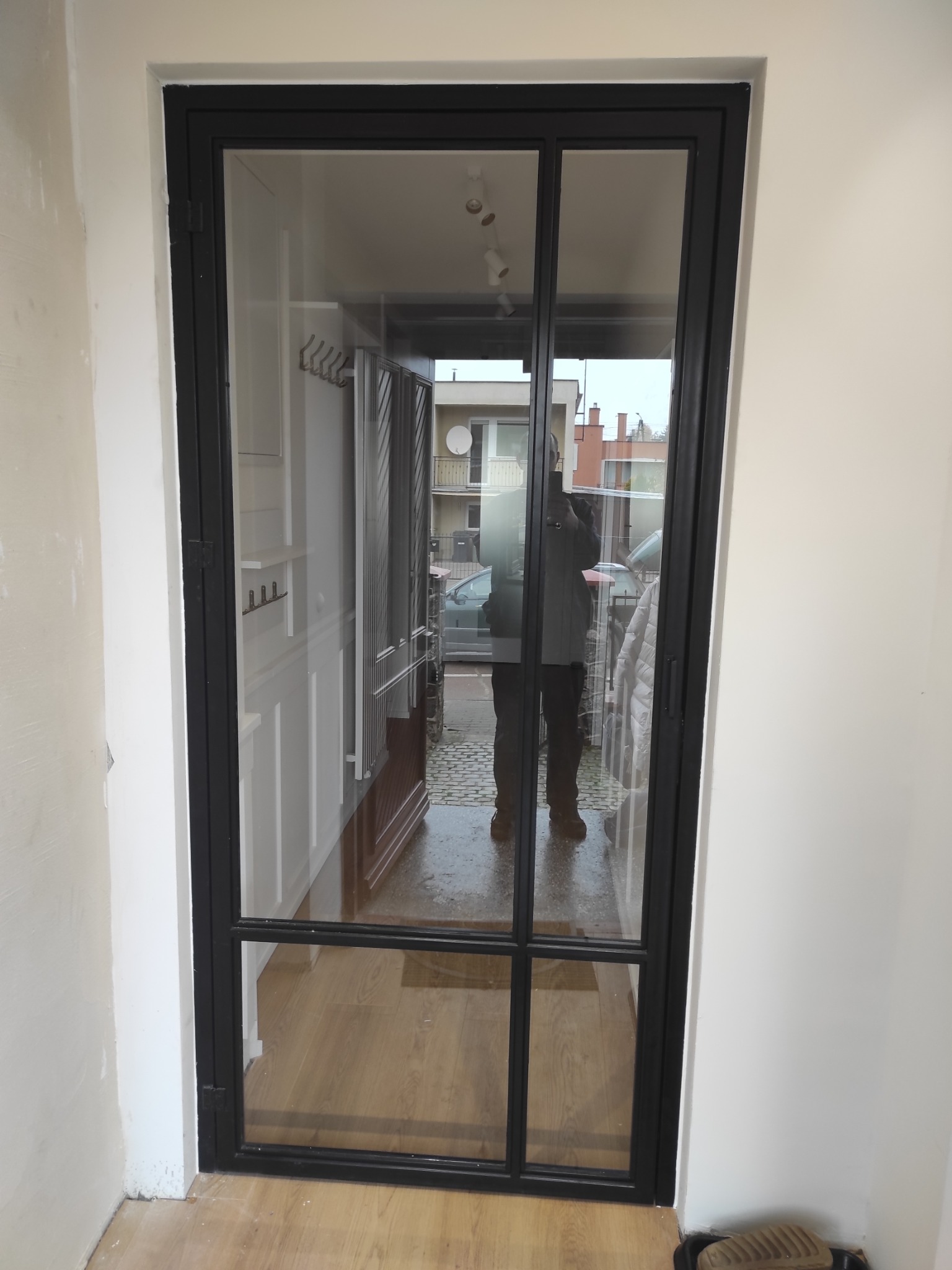 Czarna, przeszklona konstrukcja drzwiowa z podziałami, widoczna z perspektywy wnętrza, z odbiciem osoby robiącej zdjęcie na szybie. W tle widoczne elementy wyposażenia wnętrza oraz ulica.