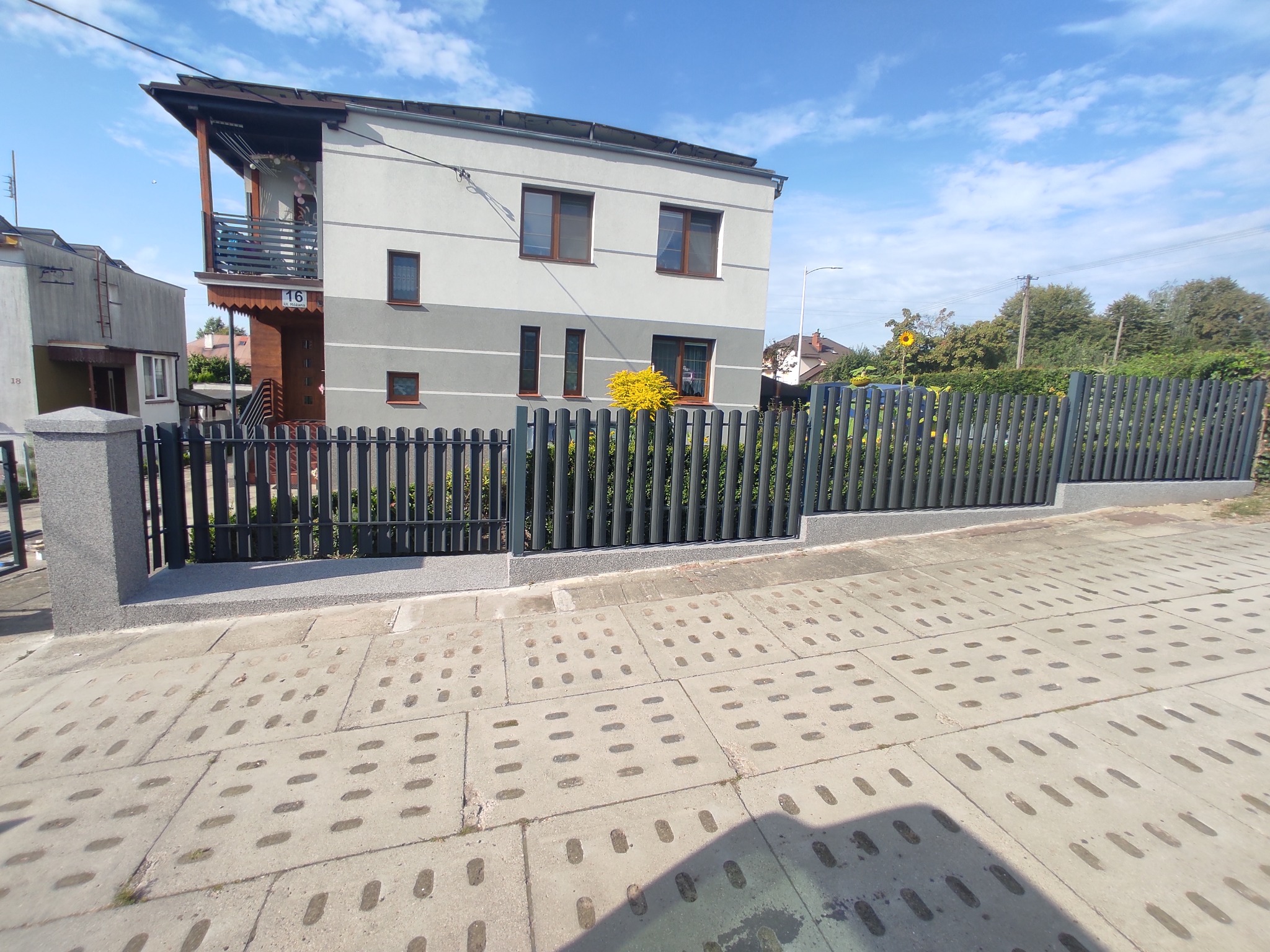 Nowoczesne ogrodzenie metalowe w kolorze grafitowym, z pionowymi sztachetami, zamontowane na niskiej podmurówce z szarego betonu, przed dwukondygnacyjnym domem z szarą elewacją i brązowymi ramami...