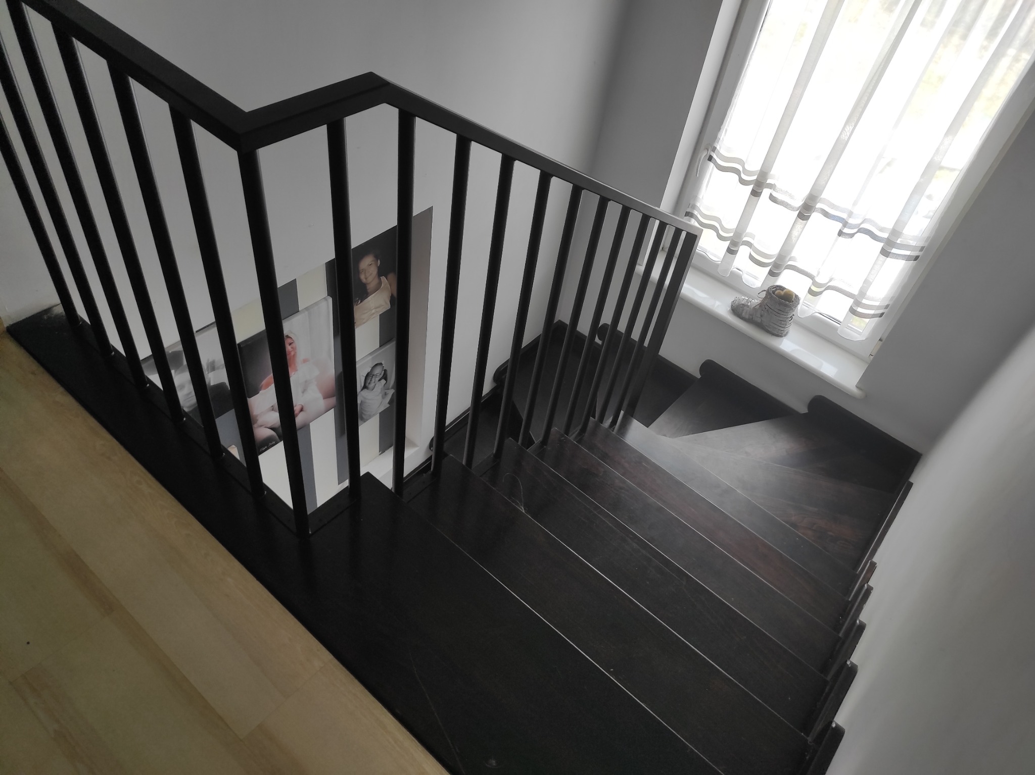 Nowoczesne, ciemne schody drewniane z czarną metalową balustradą, widok z góry, na ścianie galeria zdjęć, na parapecie okna dekoracja.
