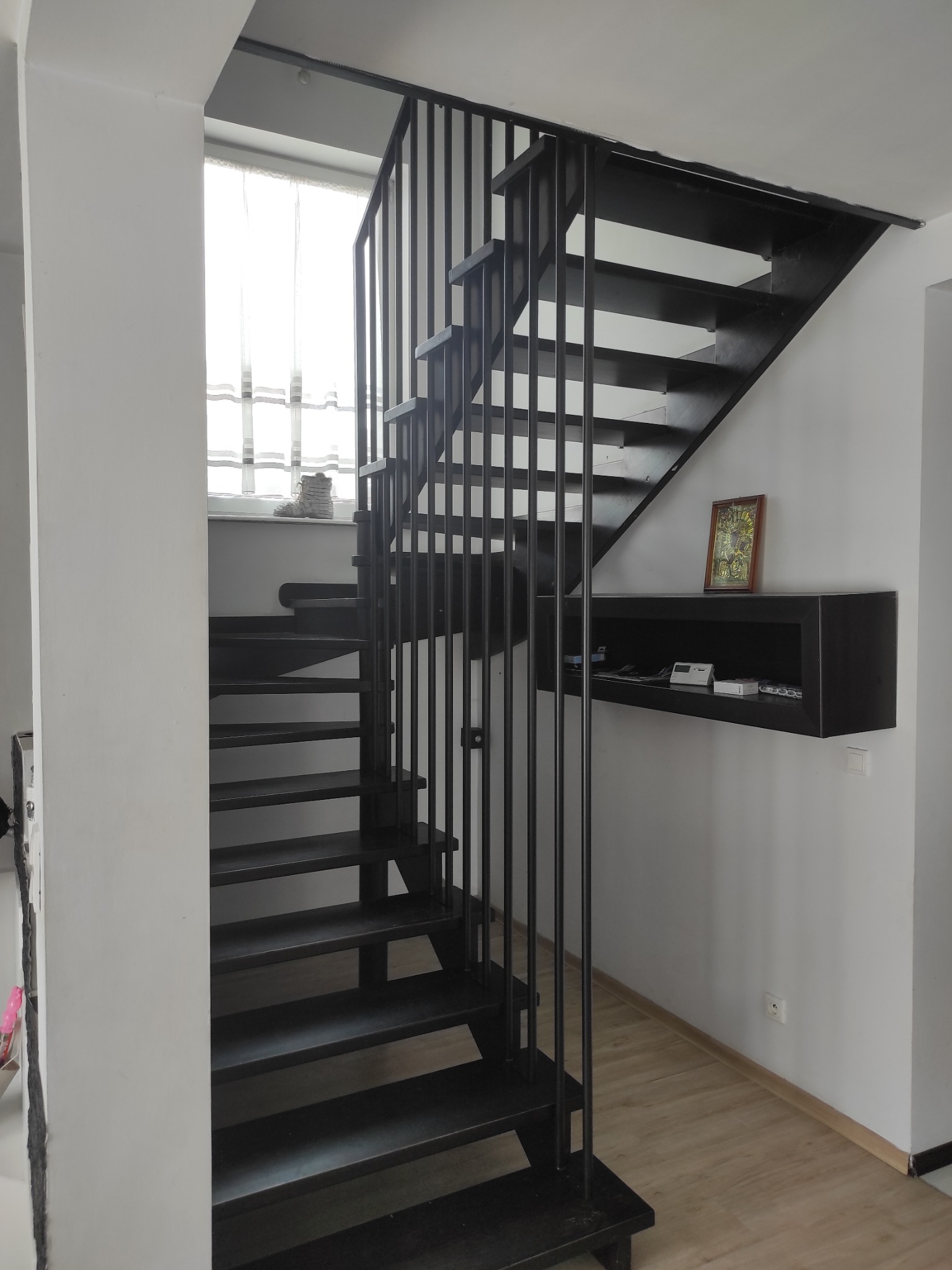 Czarne metalowe schody z pionowymi balustradami w jasnym wnętrzu, z półką na ścianie i obrazkiem w ramce.