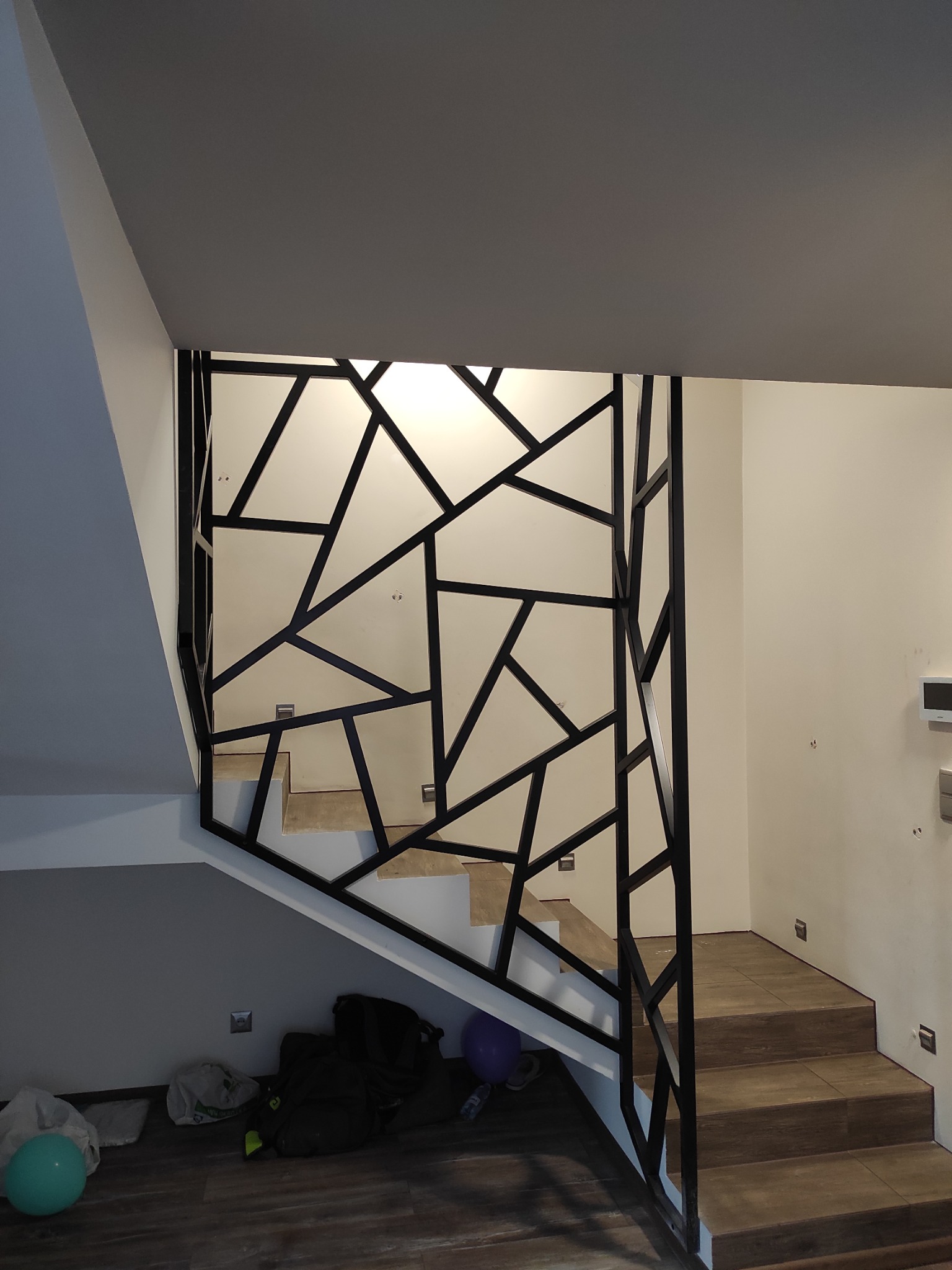 Nowoczesna balustrada schodowa z czarnego metalu o geometrycznym wzorze, widok z dołu schodów, w tle jasne ściany i drewnopodobne stopnie.