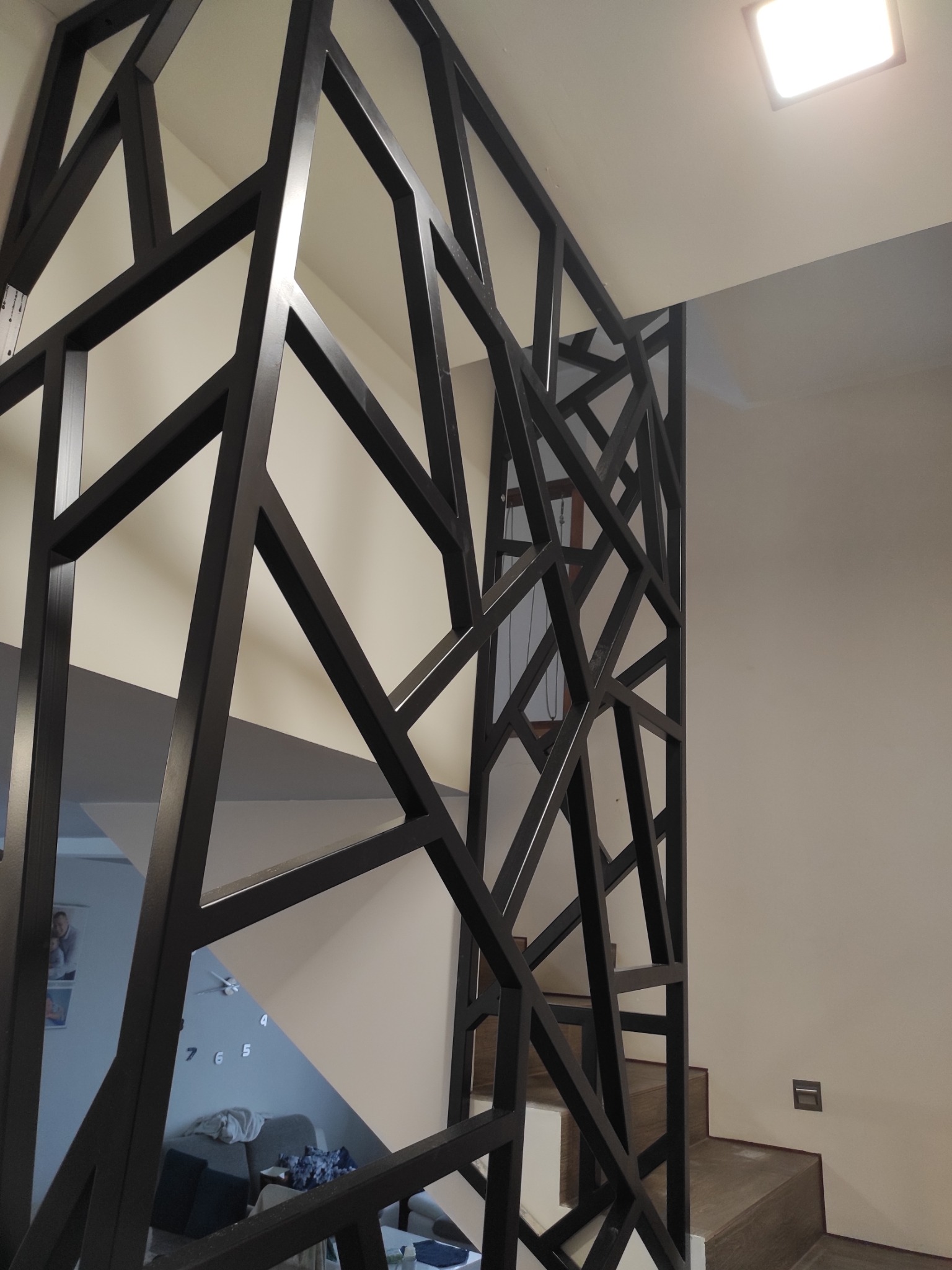 Nowoczesna, ażurowa barierka schodowa z metalu, pomalowana na czarno, z geometrycznym wzorem, widoczna na tle schodów i ściany.