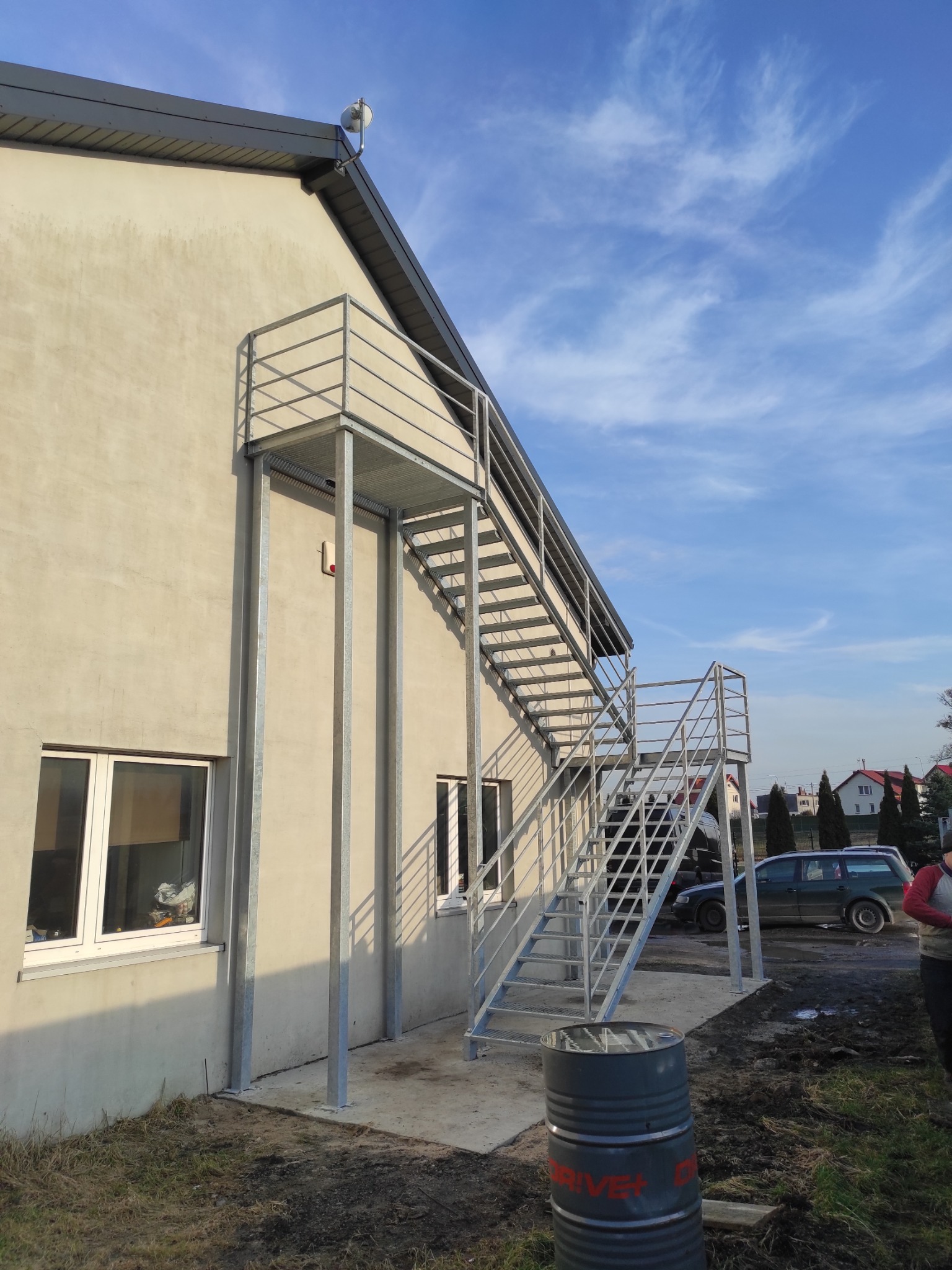 Zewnętrzne, metalowe schody ewakuacyjne przy jasnym budynku z dwoma oknami, widoczne z boku, z beczką w tle i fragmentem samochodu oraz niebem z chmurami.