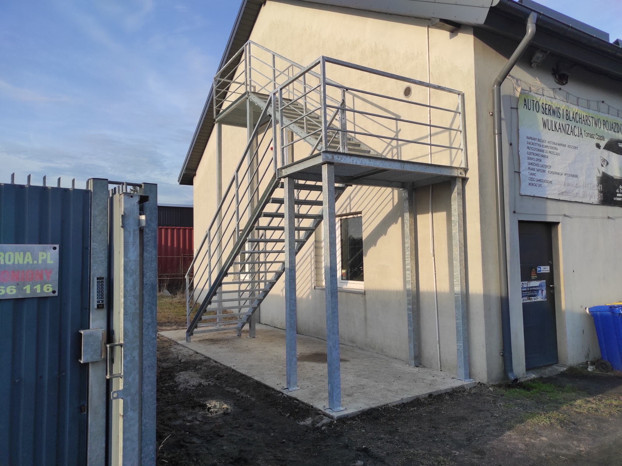 Zewnętrzne, metalowe schody z podestem prowadzące do budynku usługowego, widoczne z poziomu gruntu, z fragmentem metalowej bramy w tle.