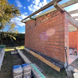 Firma Budowlana Bartłomiej Kierat - Wznoszenie ścian z czerwonej cegły dziurawki, widoczne worki z zaprawą, drewniane elementy konstrukcji dachu i szpadel, w tle błękitne niebo.