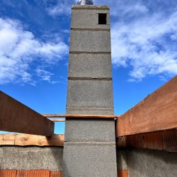 Firma Budowlana Bartłomiej Kierat - Wznoszenie komina z szarego bloczka betonowego na tle błękitnego nieba z chmurami, widoczne elementy konstrukcji więźby dachowej z drewna.