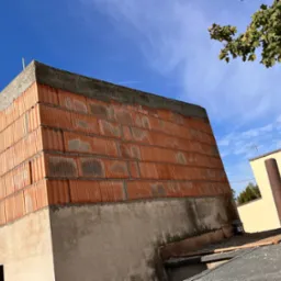 Niedokończona murowana ściana domu z betonu komórkowego, widok z dołu na tle błękitnego nieba z gałęzią drzewa w prawym górnym rogu.