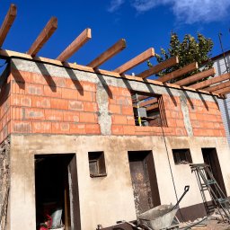 Firma Budowlana Bartłomiej Kierat - Wznoszenie dwukondygnacyjnego budynku z czerwonej cegły, widoczne otwory okienne i drzwiowe, oraz drewniana konstrukcja dachu w trakcie budowy, na pierwszym planie taczka i drabina.