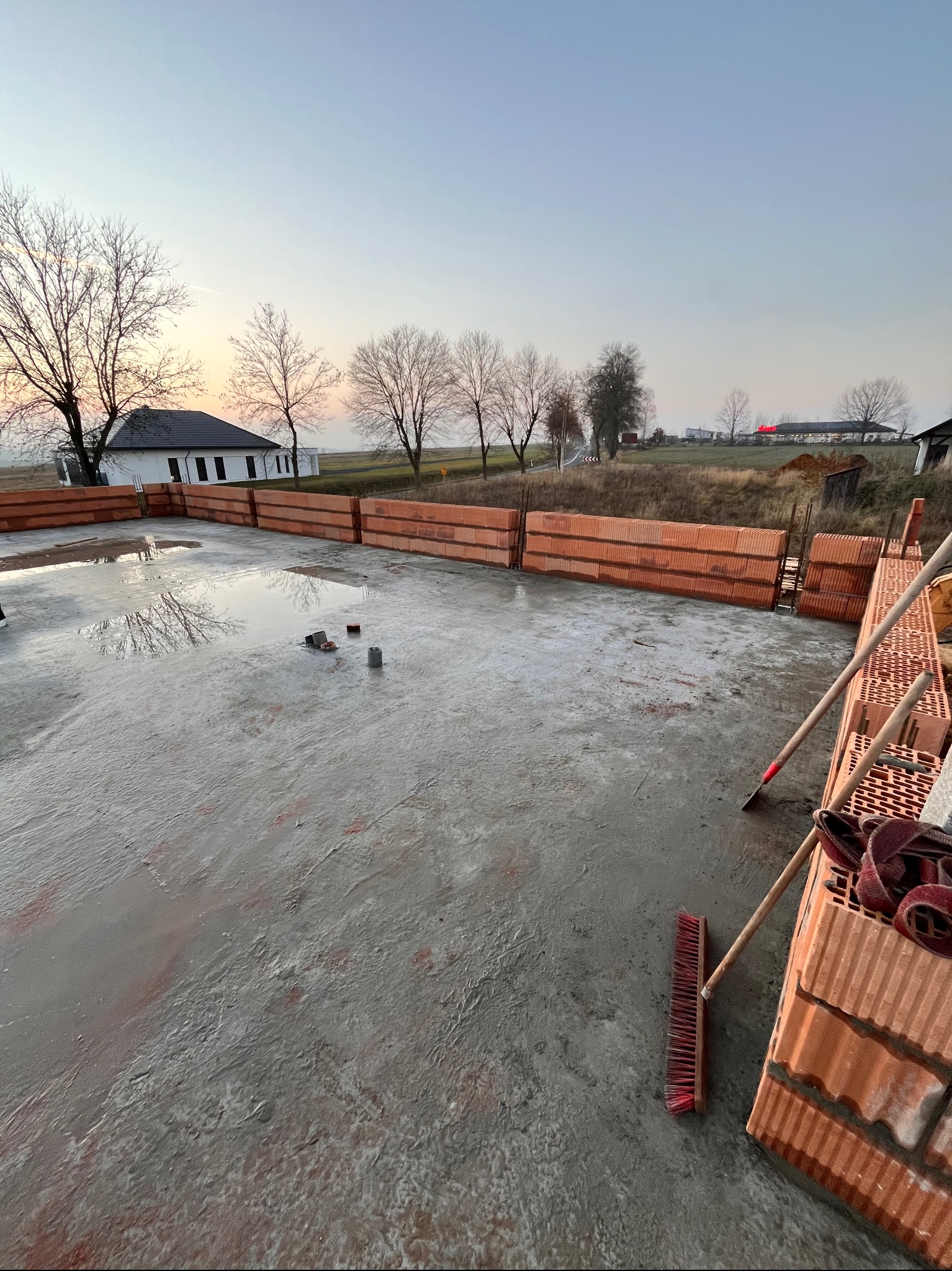 Wylany fundament z częściowo wymurowanymi ścianami z pustaków ceramicznych, widoczne narzędzia murarskie, w tle budynek jednorodzinny i drzewa o zachodzie słońca.