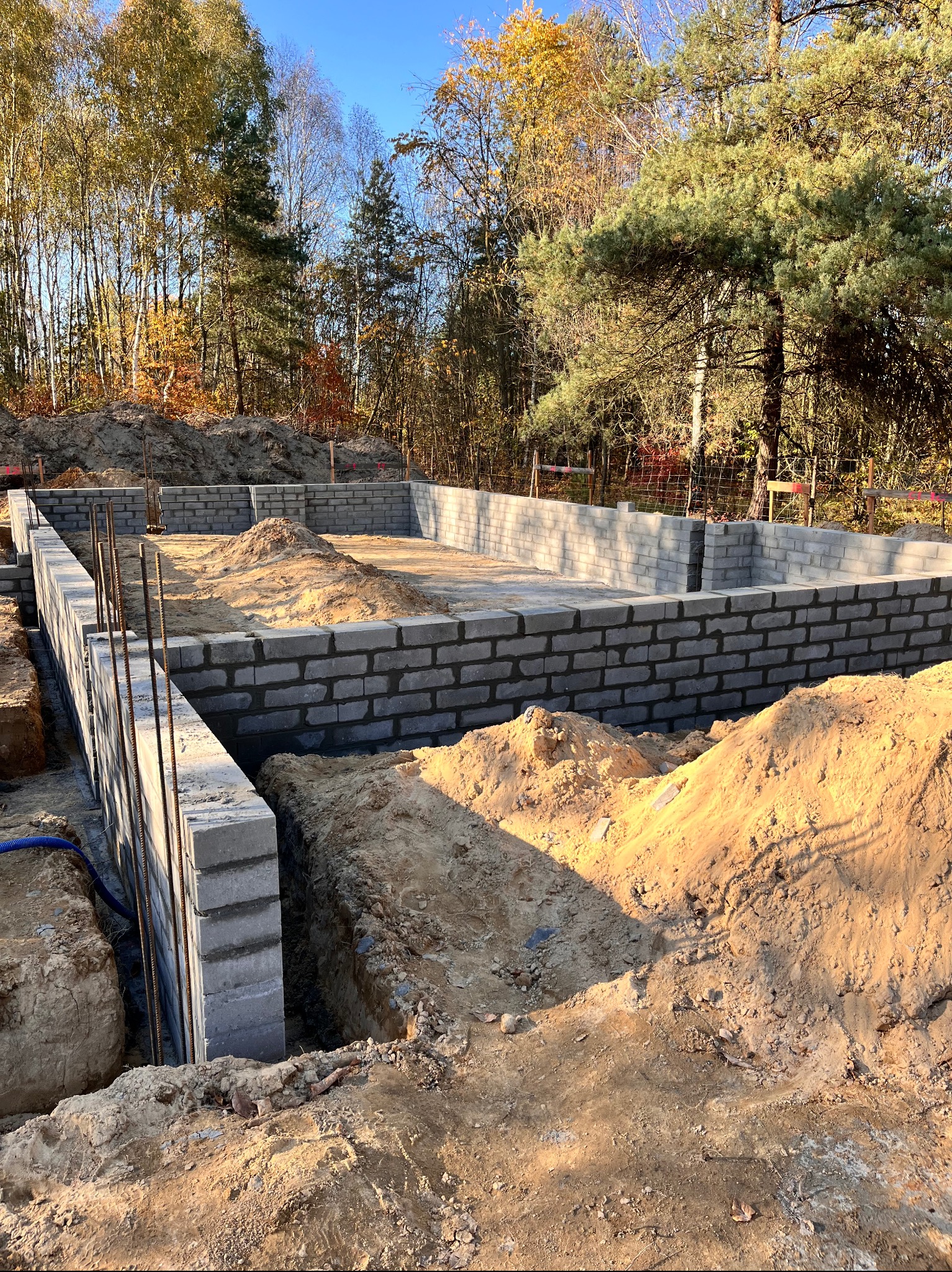 Wznoszenie fundamentów z bloczków betonowych na działce z drzewami w tle, widoczne wystające pręty zbrojeniowe i nasypana ziemia.