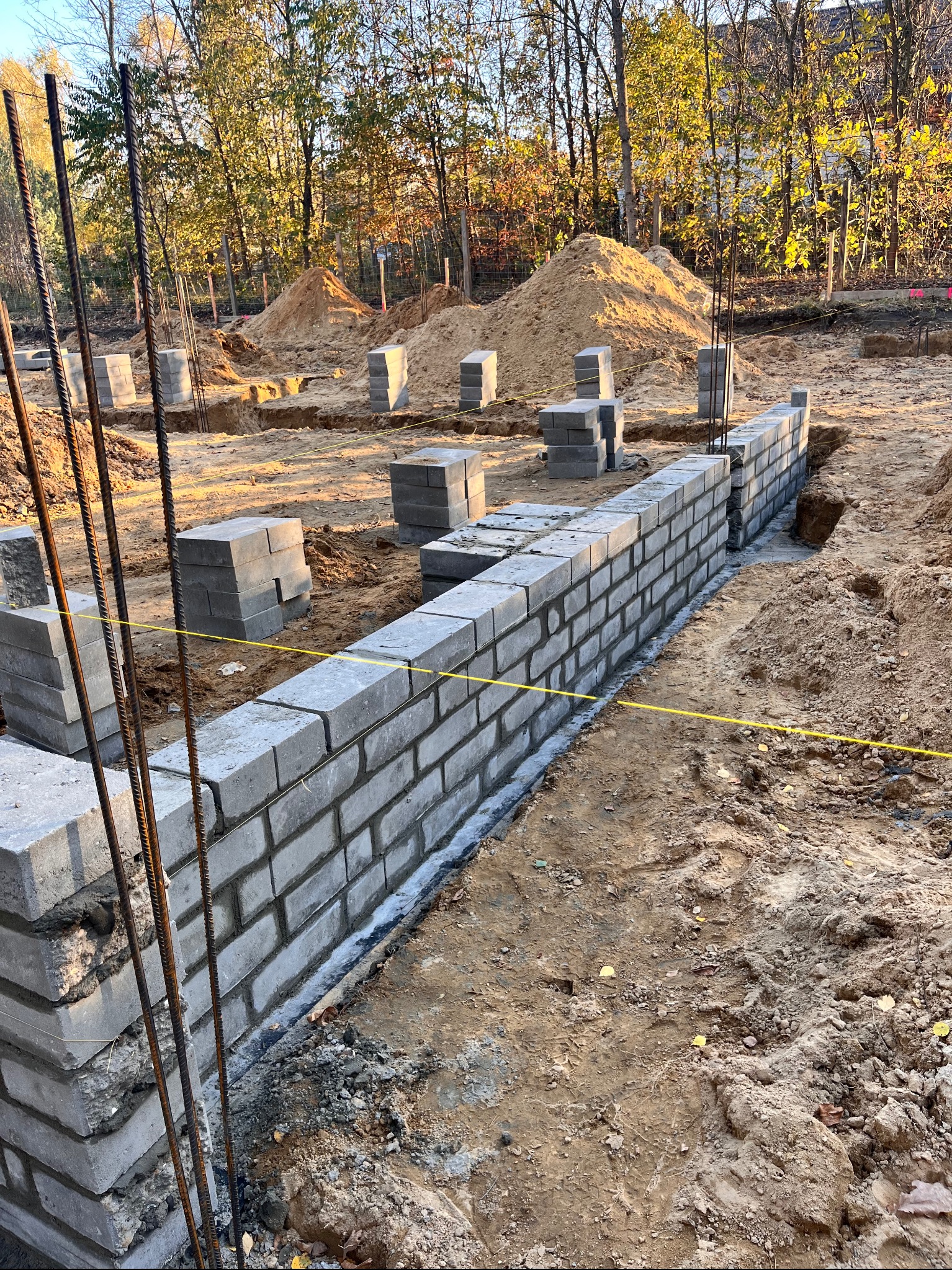 Wznoszenie ściany fundamentowej z betonowych bloczków, widoczne wystające pręty zbrojeniowe i żółta linka wyznaczająca linię, w tle hałdy piachu i drzewa.