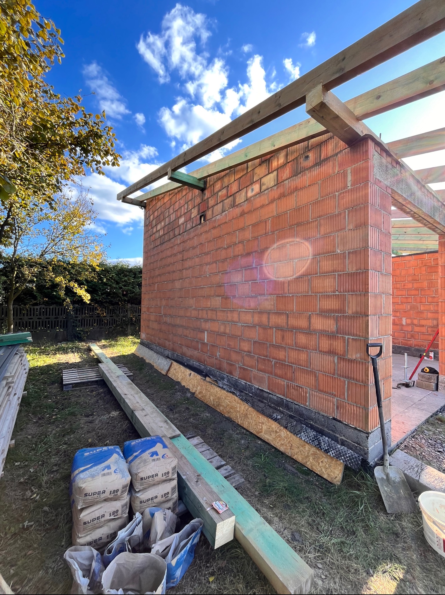 Wznoszenie ścian z czerwonej cegły dziurawki, widoczne worki z zaprawą, drewniane elementy konstrukcji dachu i szpadel, w tle błękitne niebo.