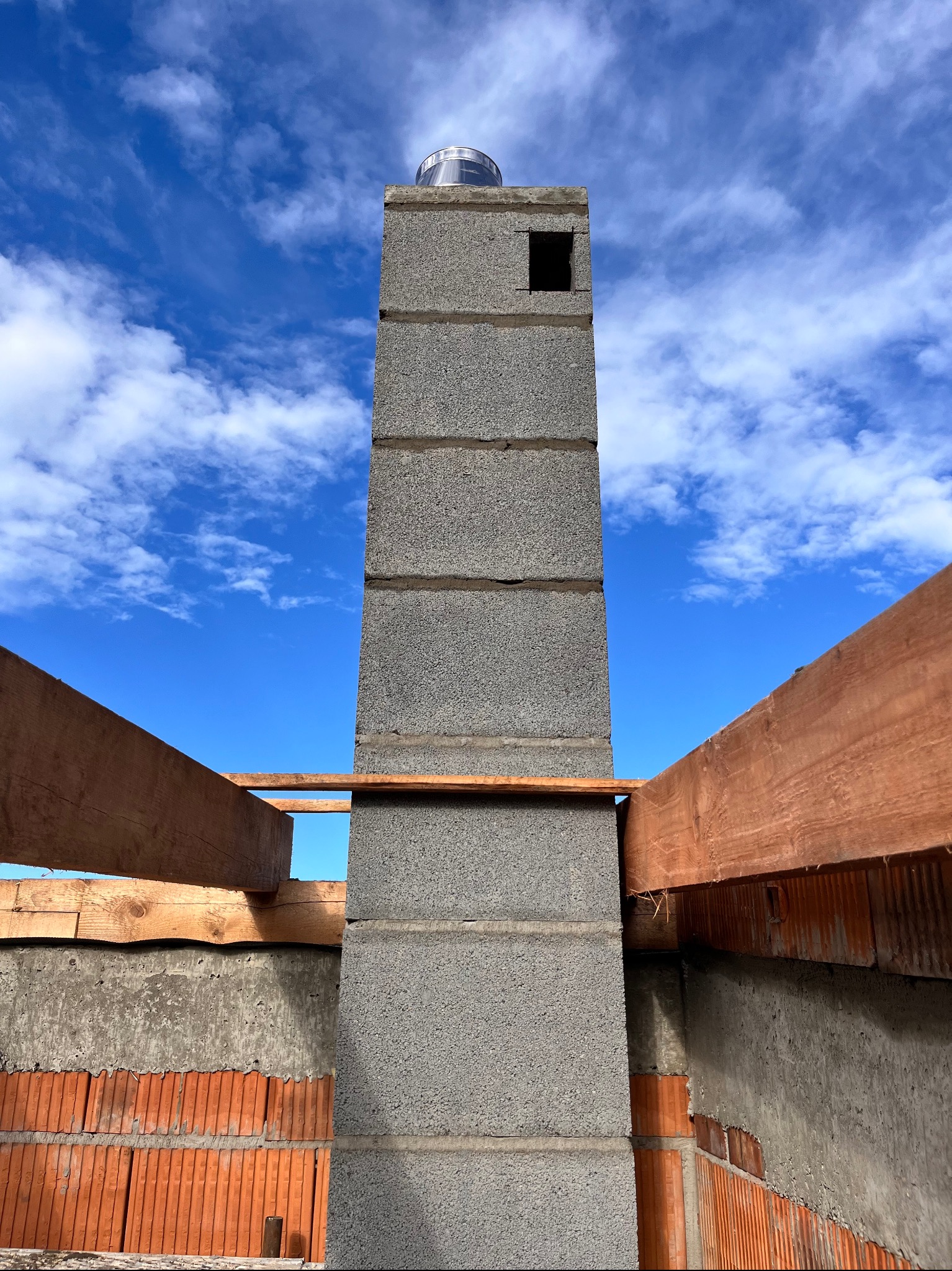 Wznoszenie komina z szarego bloczka betonowego na tle błękitnego nieba z chmurami, widoczne elementy konstrukcji więźby dachowej z drewna.