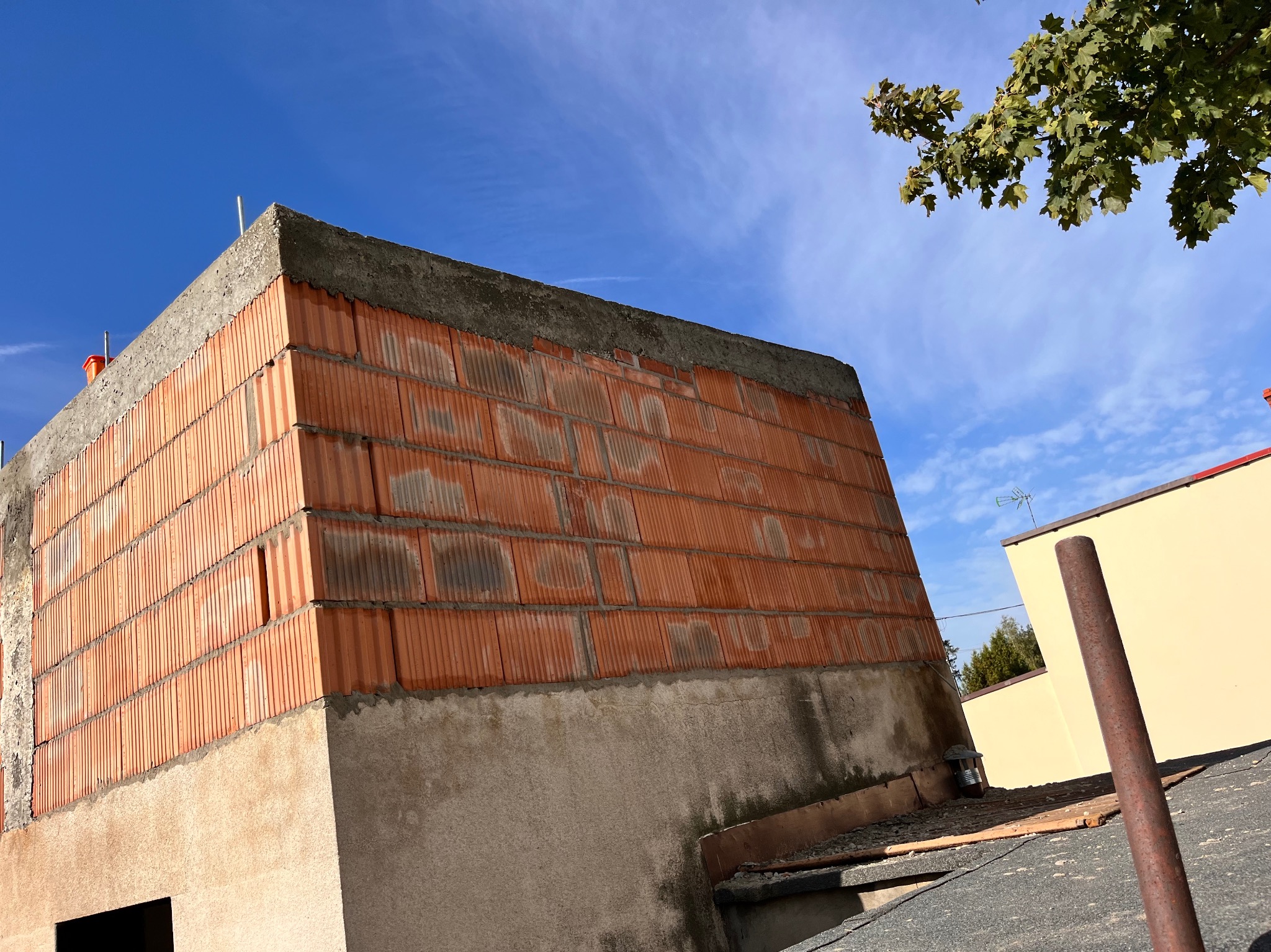 Niedokończona murowana ściana domu z betonu komórkowego, widok z dołu na tle błękitnego nieba z gałęzią drzewa w prawym górnym rogu.