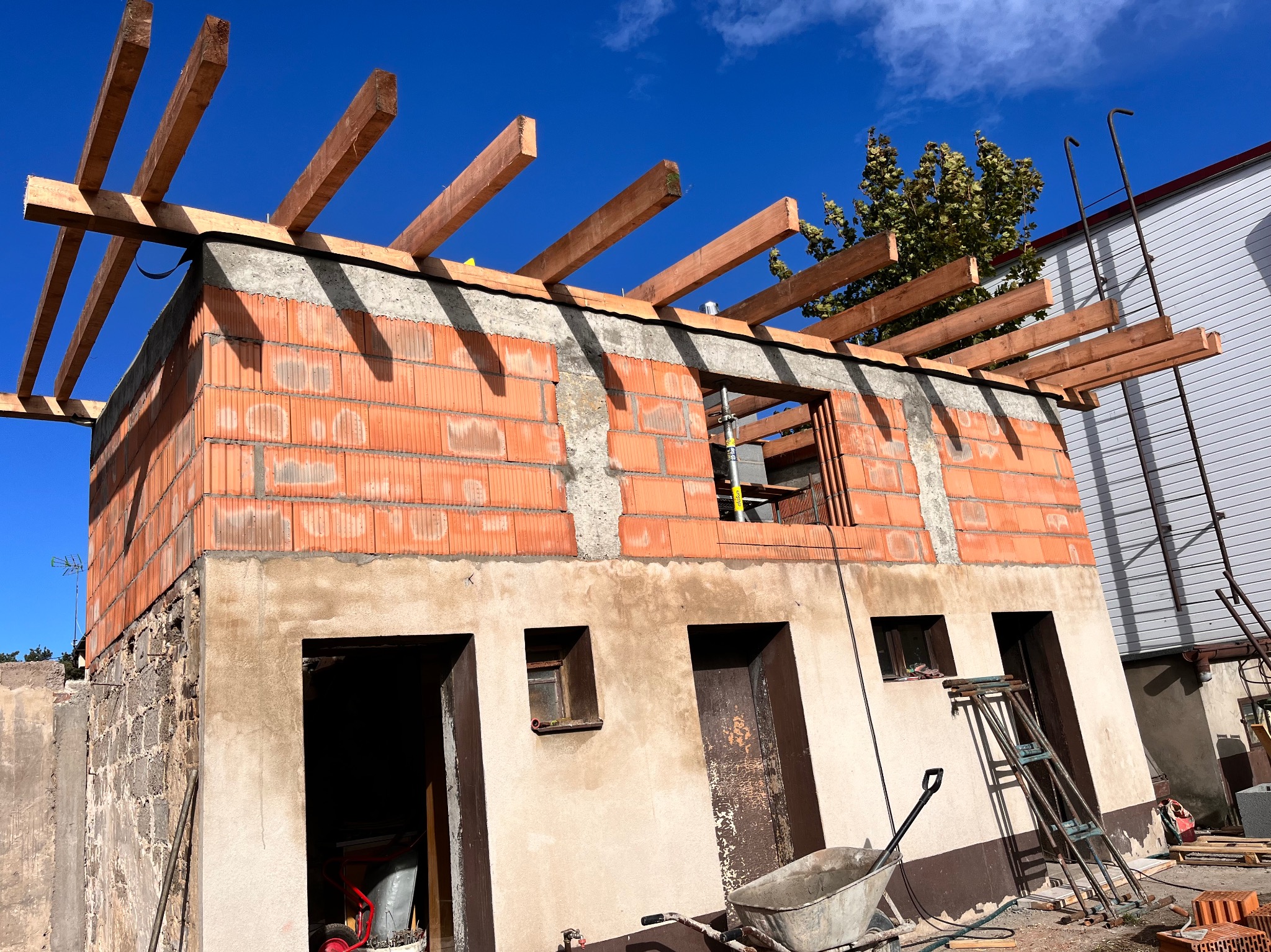 Wznoszenie dwukondygnacyjnego budynku z czerwonej cegły, widoczne otwory okienne i drzwiowe, oraz drewniana konstrukcja dachu w trakcie budowy, na pierwszym planie taczka i drabina.