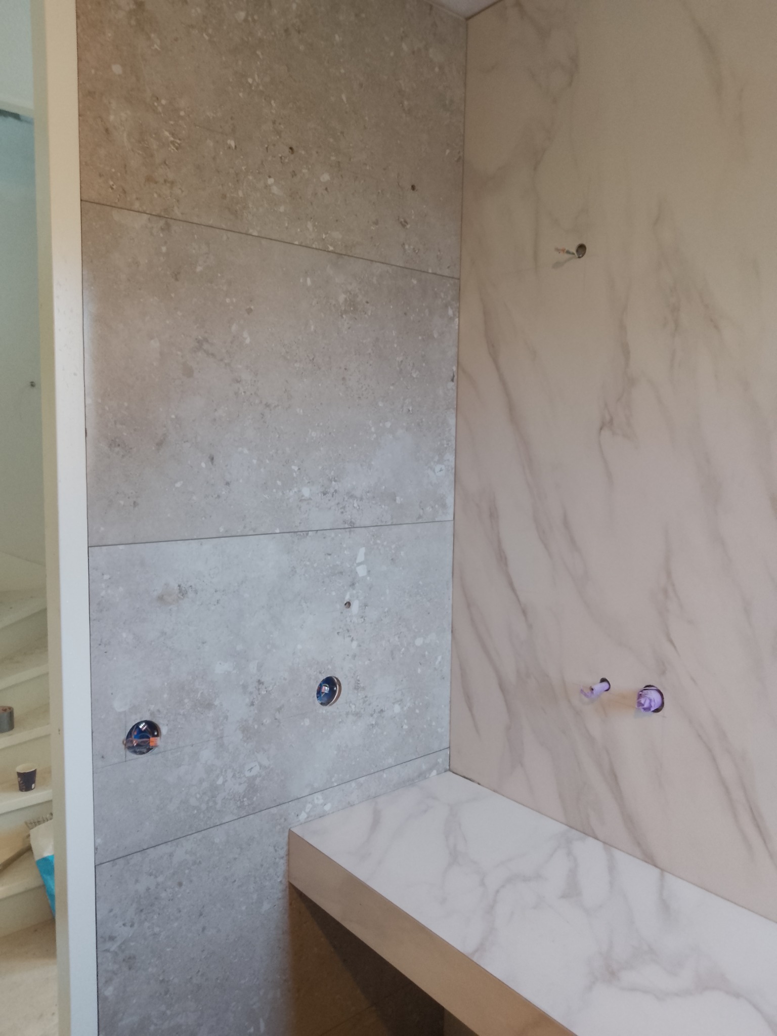 Fragment łazienki w trakcie wykańczania: ściana wyłożona płytkami imitującymi kamień w odcieniach szarości, z widocznymi otworami na instalacje, kontrastująca z nią ściana wyłożona płytkami...