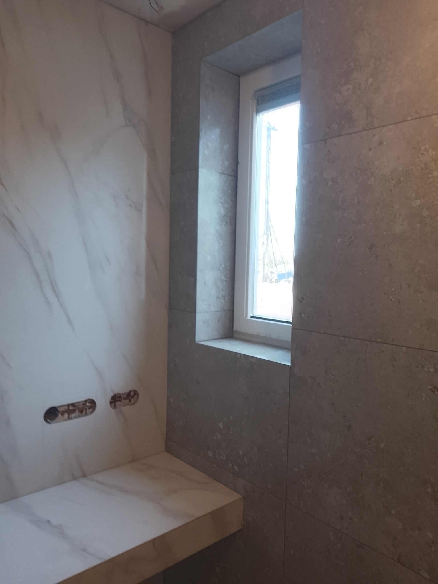 Ściana łazienki wyłożona płytkami imitującymi marmur i kamień, z widocznymi otworami na instalację wodną i parapetem pod oknem.