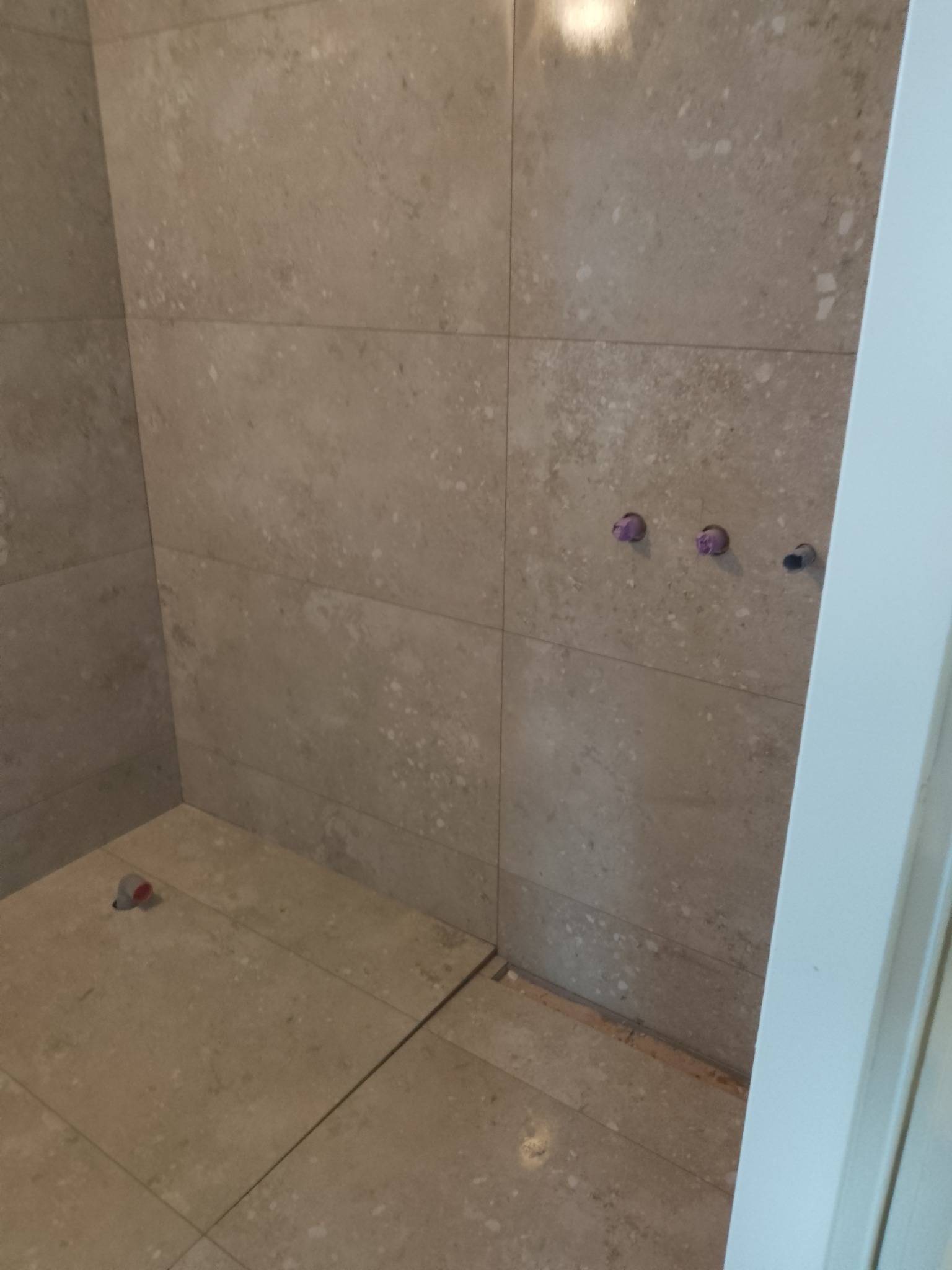 Świeżo ułożone, beżowe płytki podłogowe i ścienne w łazience w Gdowie, z widocznymi trzema otworami na baterię i rurę odpływową zabezpieczoną zatyczką.