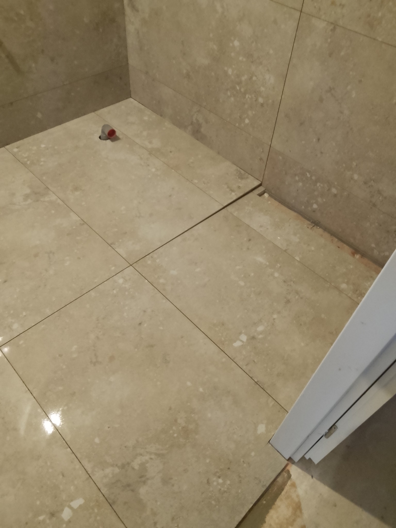 Fragment podłogi i ścian w łazience wyłożonych jasnymi, beżowymi płytkami imitującymi kamień. Widoczny odpływ podłogowy z czerwoną zatyczką oraz biała ościeżnica drzwiowa. Fuga między płytkami jest...