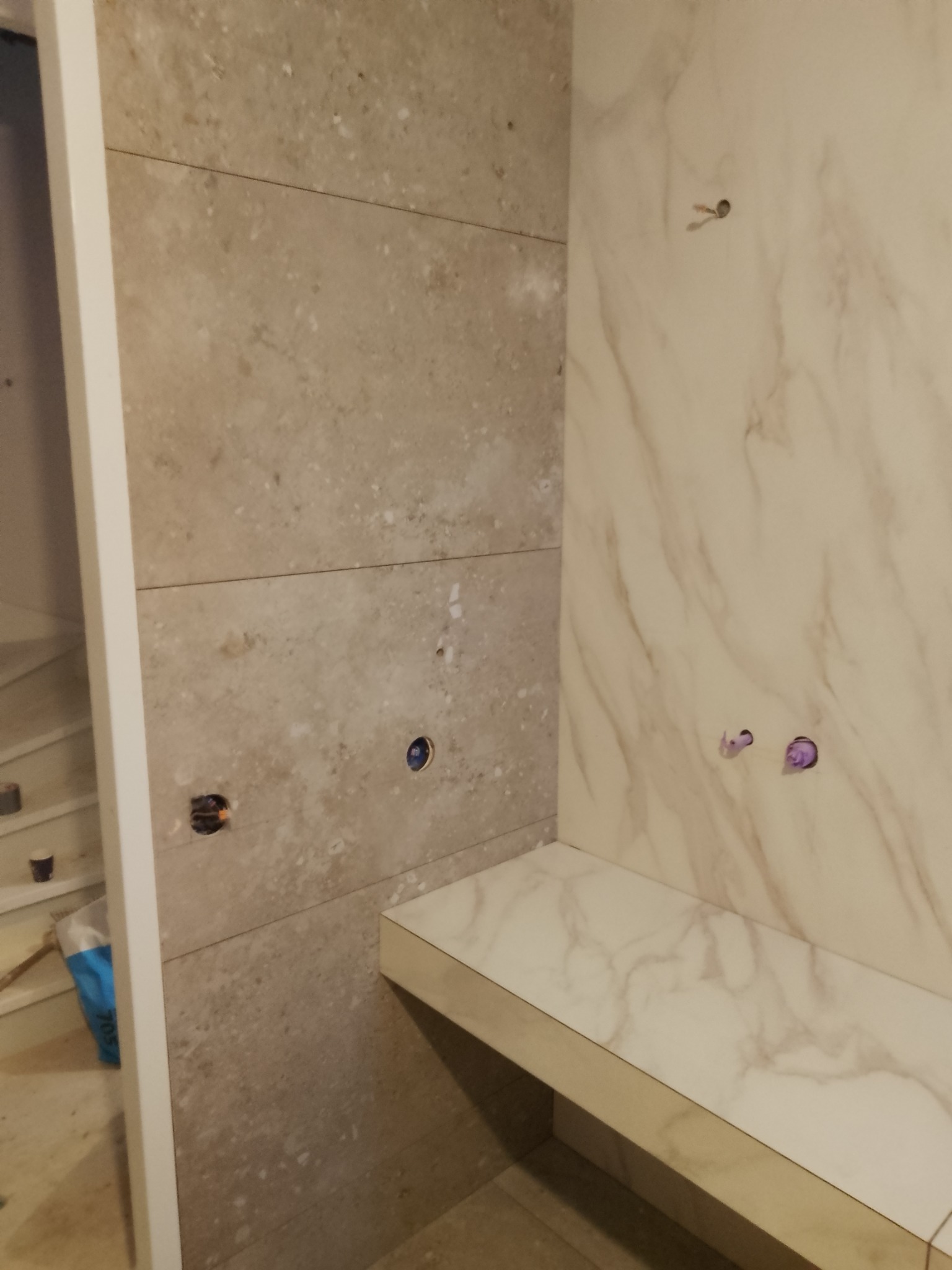 Ściana łazienki wyłożona płytkami imitującymi kamień, z widocznymi otworami na instalacje i półką z płytek marmurowych, w trakcie prac wykończeniowych.