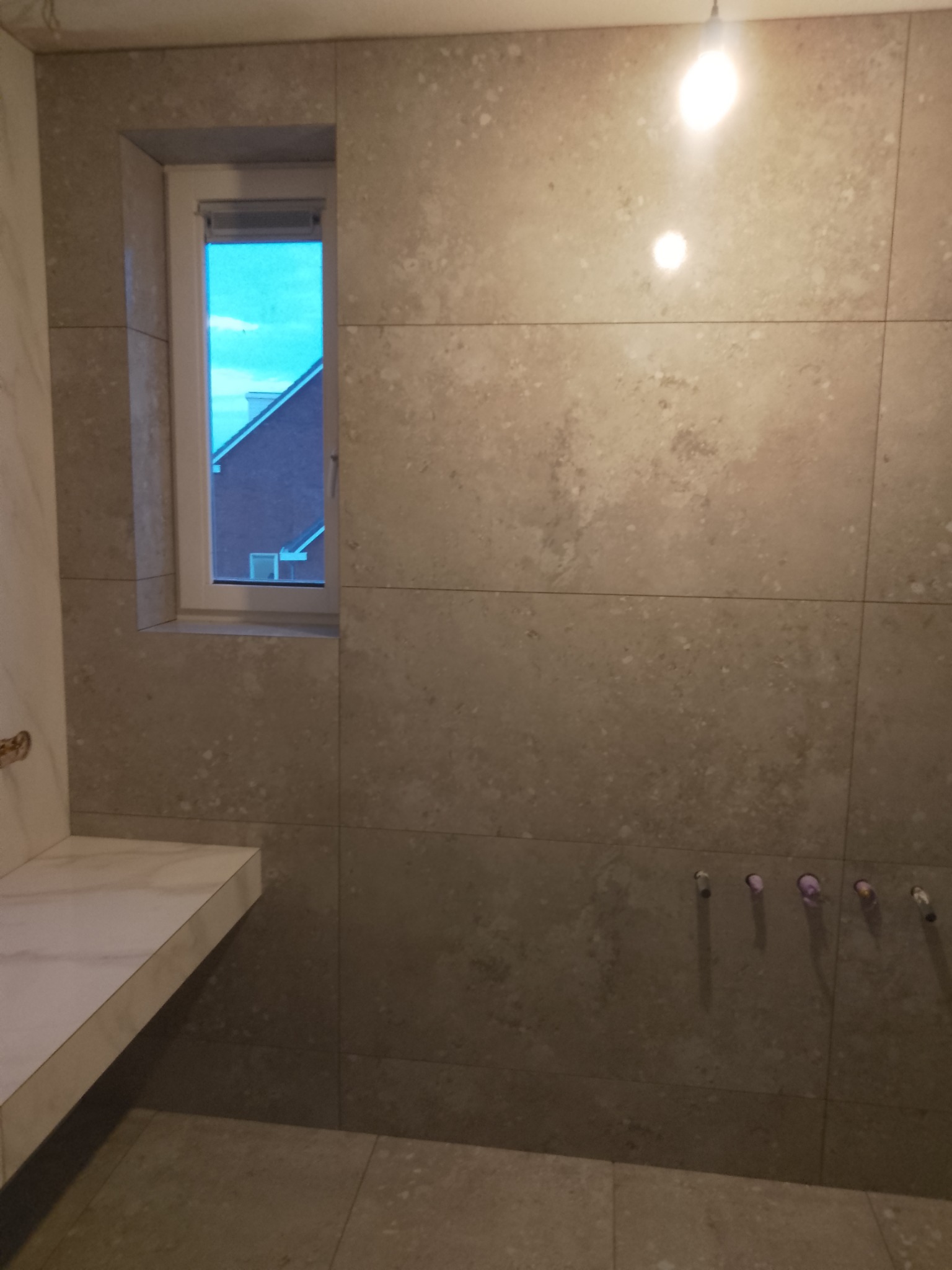 Ściana w łazience wyłożona beżowymi płytkami imitującymi kamień, z oknem i wystającym blatem umywalkowym z jasnego marmuru; widoczne wystające rurki instalacji wodnej.