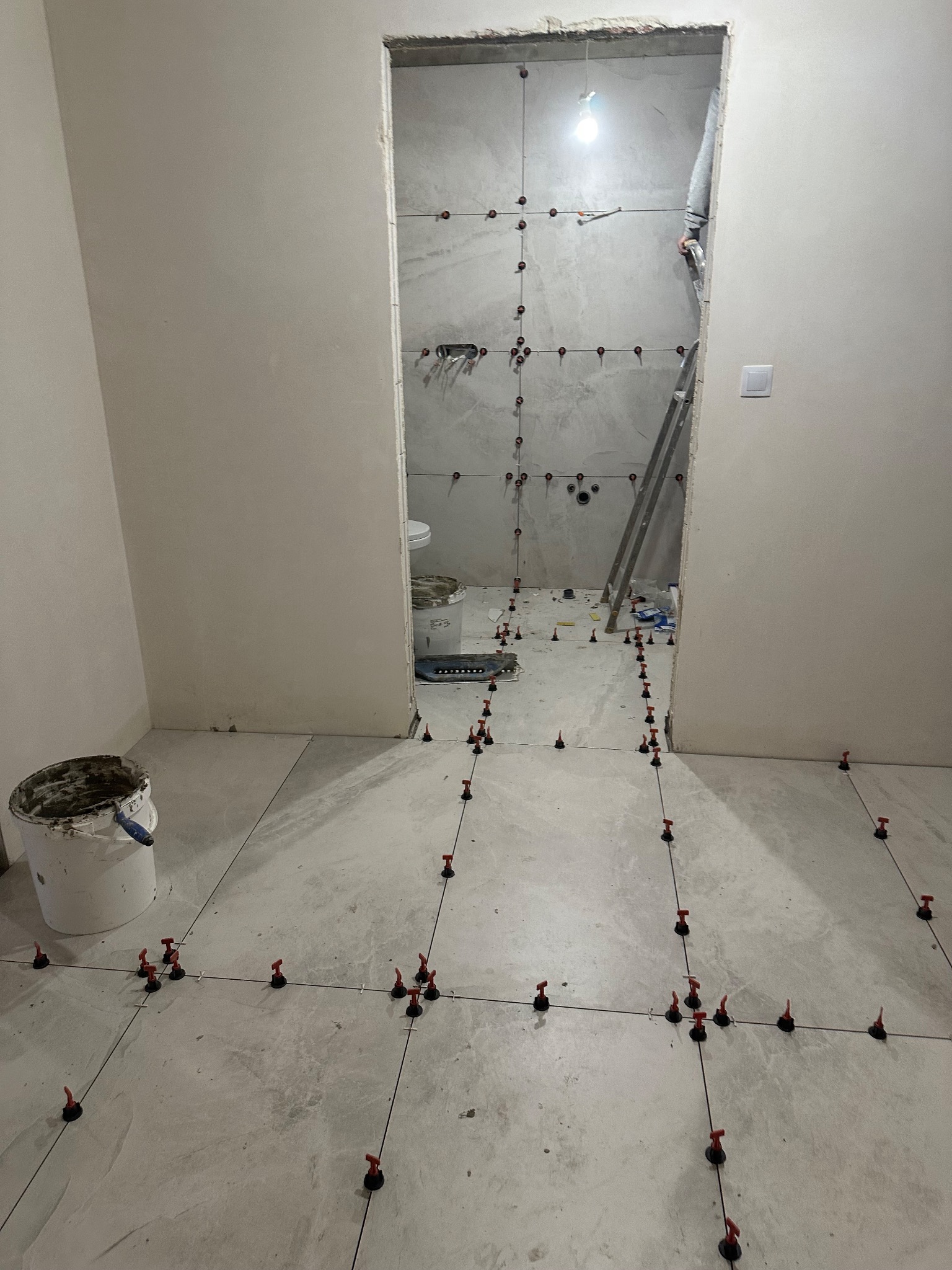 Układanie płytek podłogowych w łazience z użyciem systemu poziomowania. Widoczny fragment ściany z ułożonymi płytkami i drabina. Wiadro z klejem w rogu pomieszczenia.