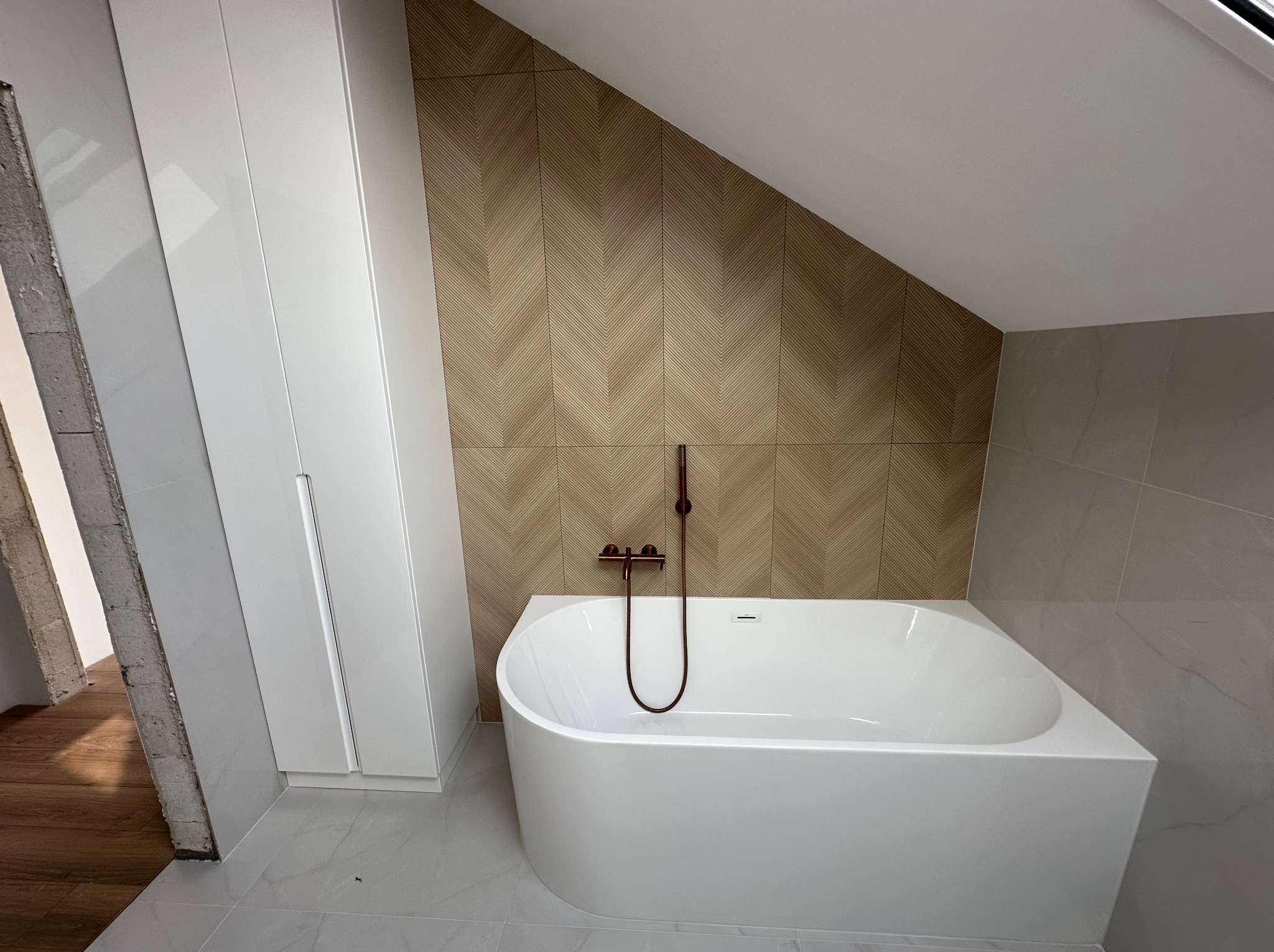 Nowoczesna łazienka na poddaszu z białą wanną, drewnopodobnymi płytkami ułożonymi w jodełkę i minimalistyczną armaturą w kolorze miedzi. Widoczna zabudowa szafy.