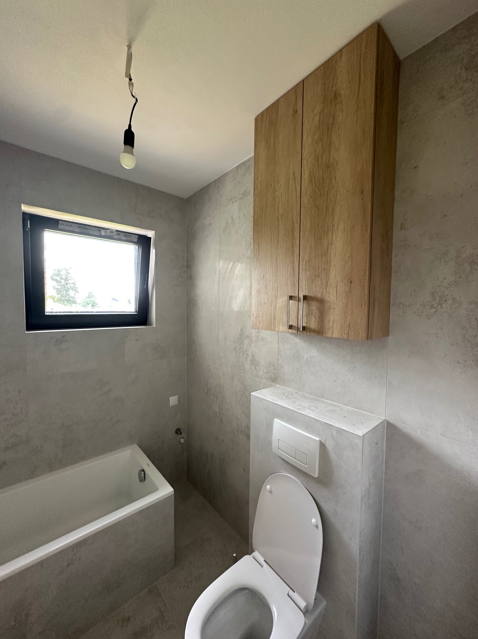 Wykończona łazienka w szarościach z drewnianą szafką wiszącą nad toaletą. Widoczna wanna, okno i surowa żarówka. Minimalistyczny design.