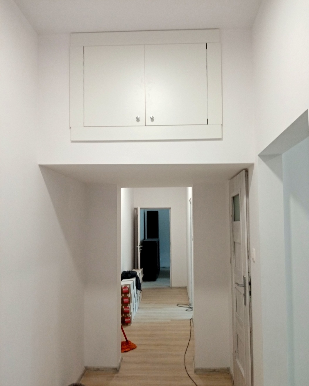 Wnętrze korytarza po odświeżeniu: białe ściany, zabudowa szafek pod sufitem z chromowanymi uchwytami, panele podłogowe imitujące drewno, widok na dalszą część mieszkania.